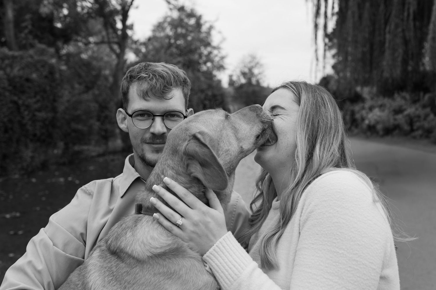 A couple laughs together with their dog in a black and white outdoor setting.