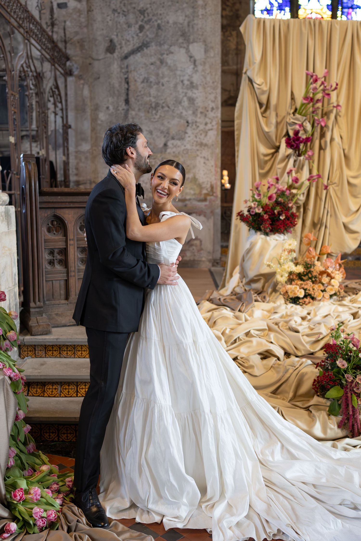 A couple shares an intimate moment in formal wedding attire against an elegant backdrop with draped fabric and floral decor.