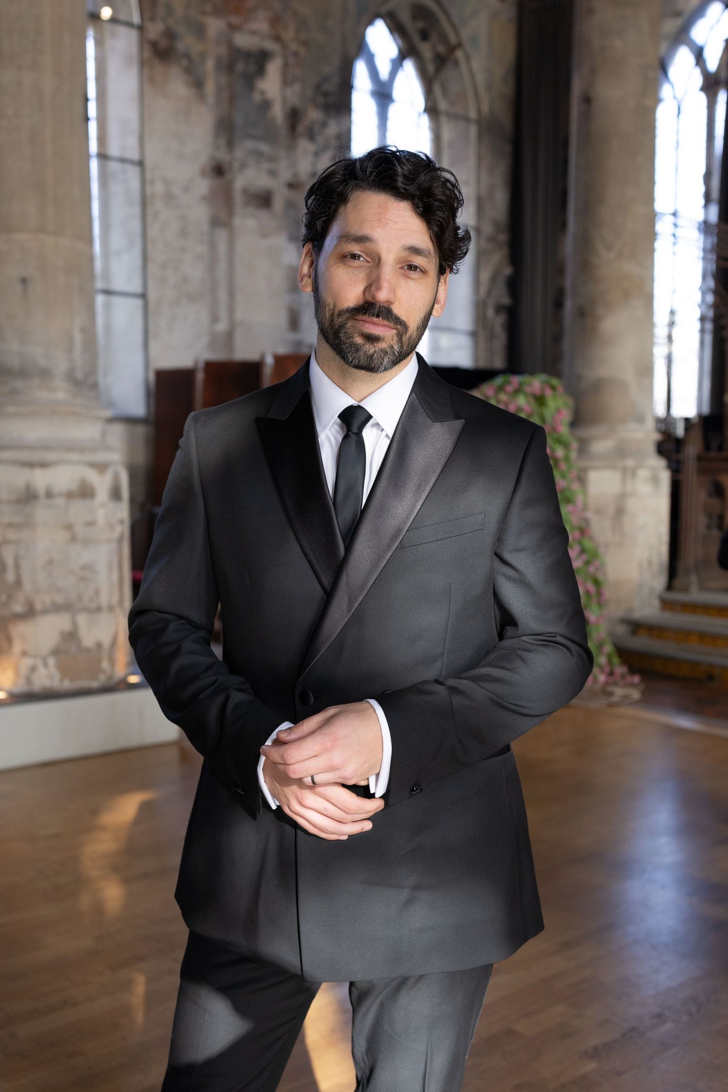 A groom in a black suit gestures while standing in a historic building with arched windows and stone columns.