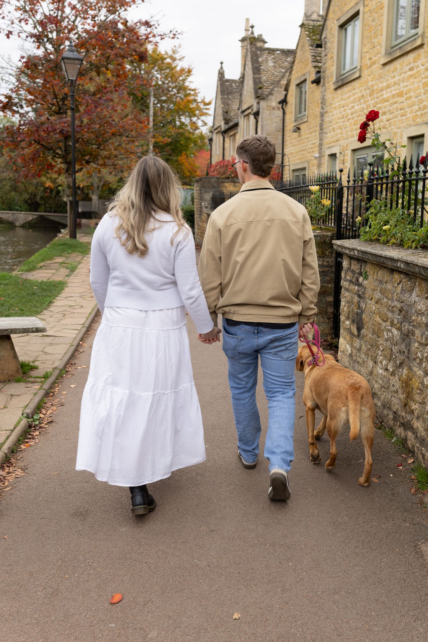 A couple walks their brown dog along a path beside stone buildings on an autumn day.