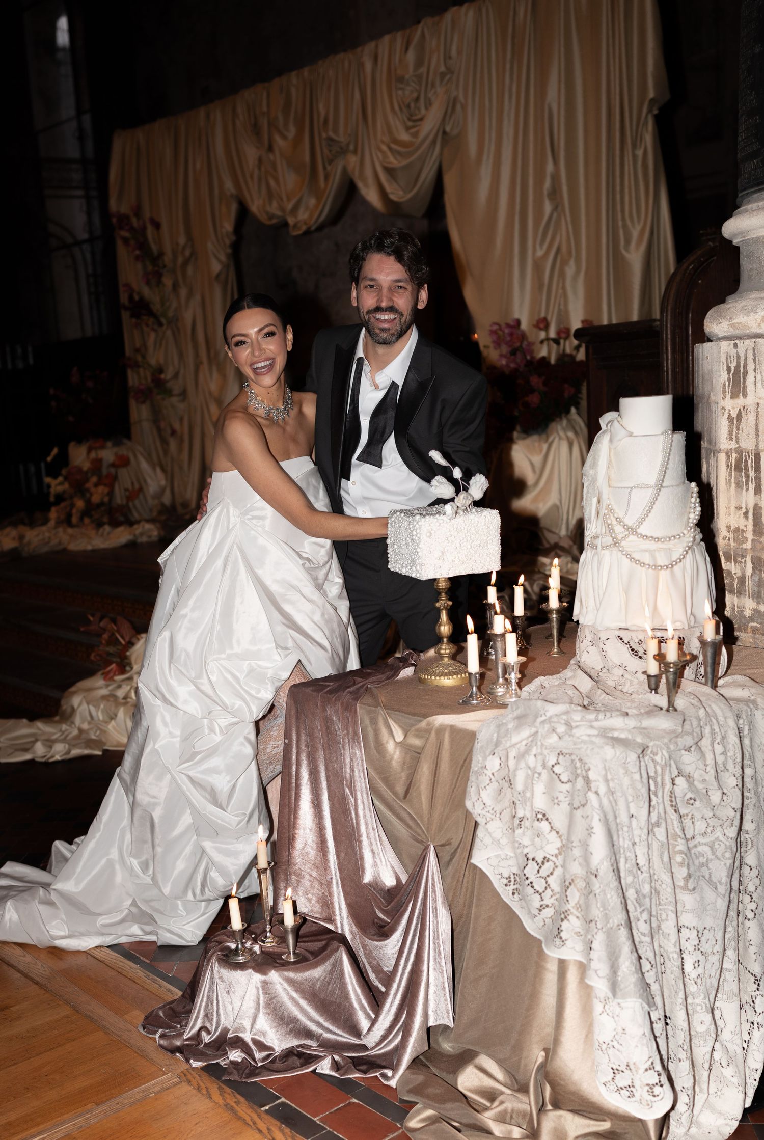 A couple cuts their elegant white wedding cake at a reception decorated with champagne-colored satin tablecloths and candles.