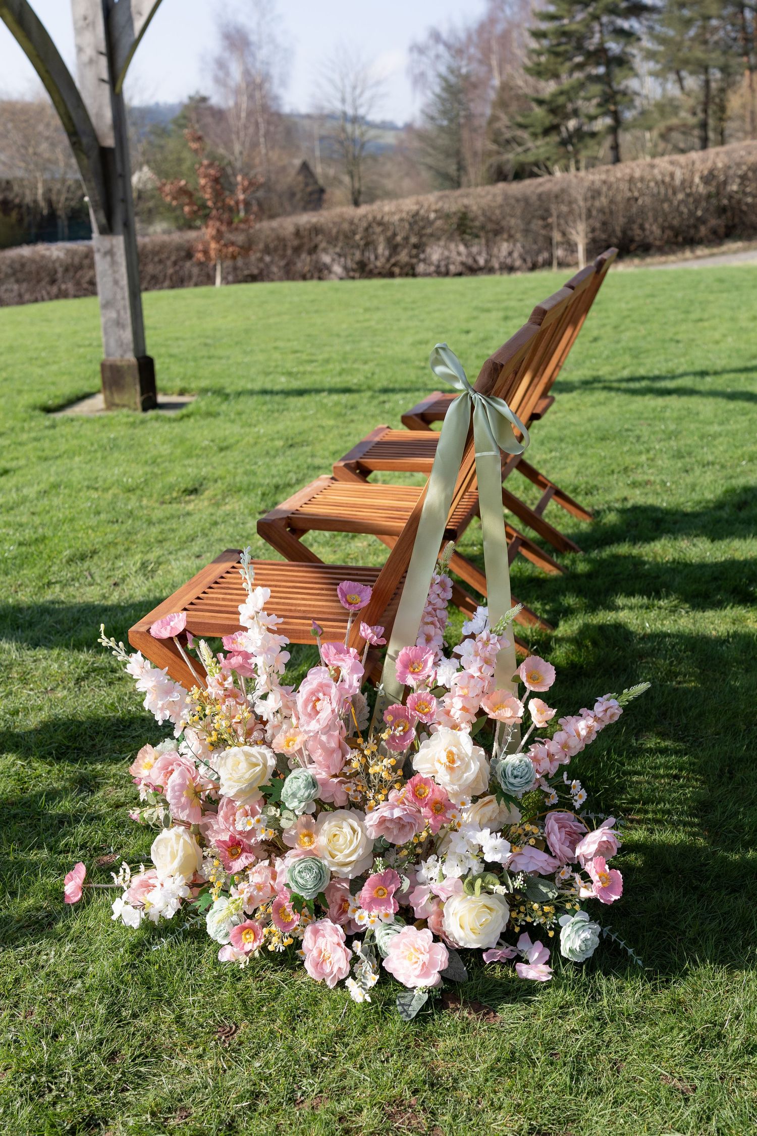 Outdoor wedding ceremony setup with wooden folding chairs adorned with sage ribbon and pink floral arrangements on green grass.