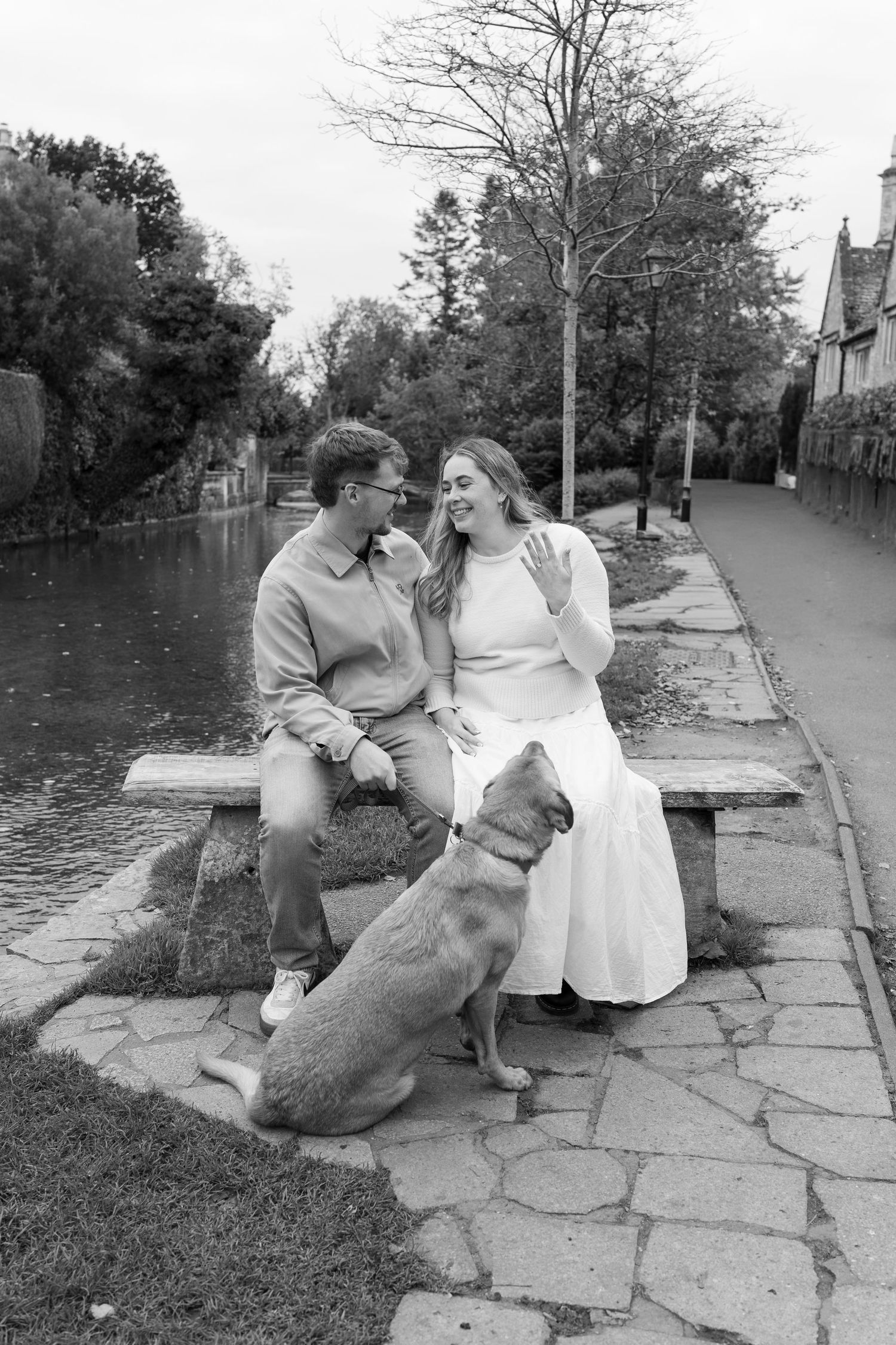 A couple sits with their dog on stone steps by a river in black and white.