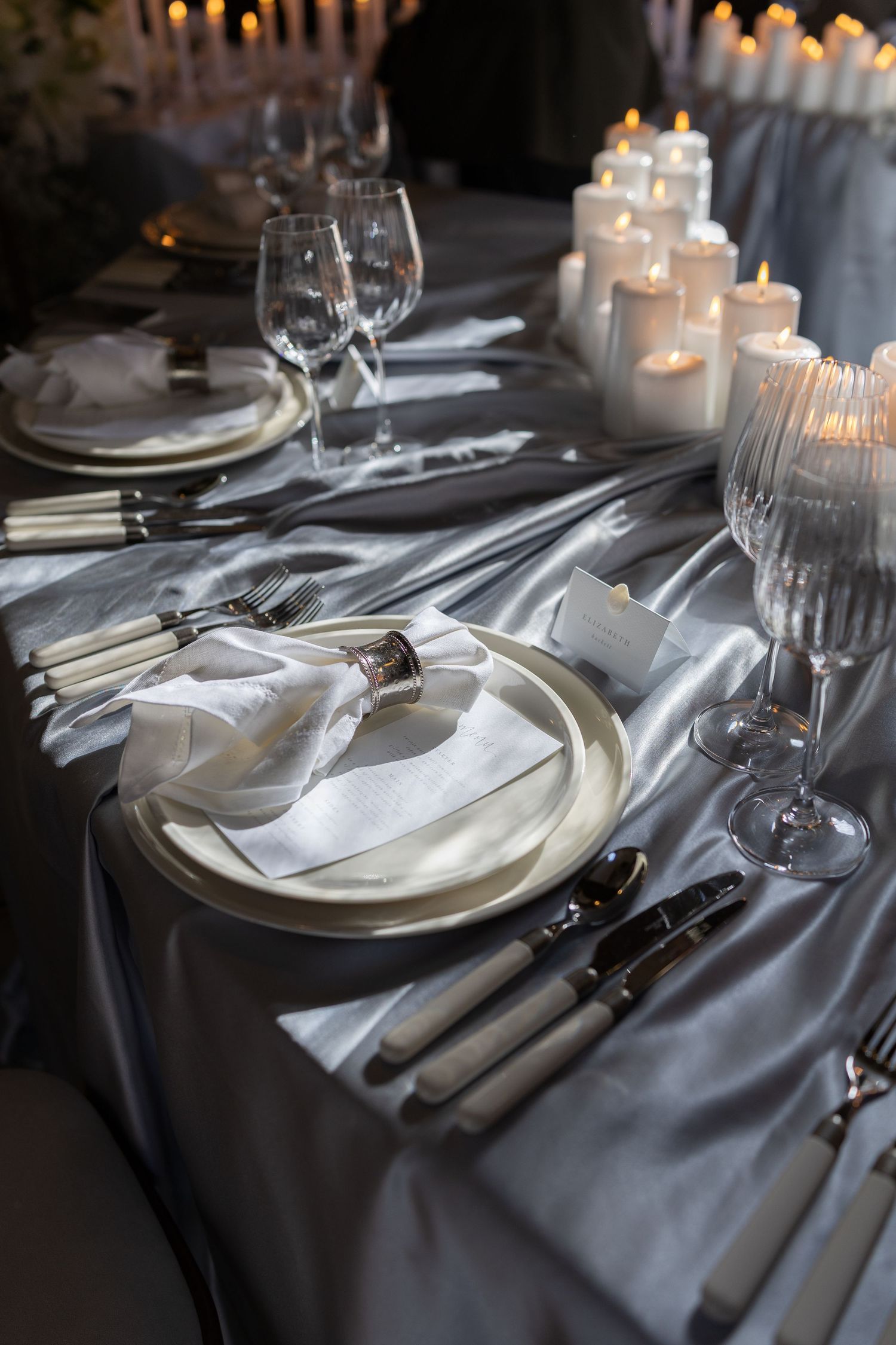 Formal wedding  table setting with silver cutlery, white plates, and glowing candles on blue satin tablecloth.