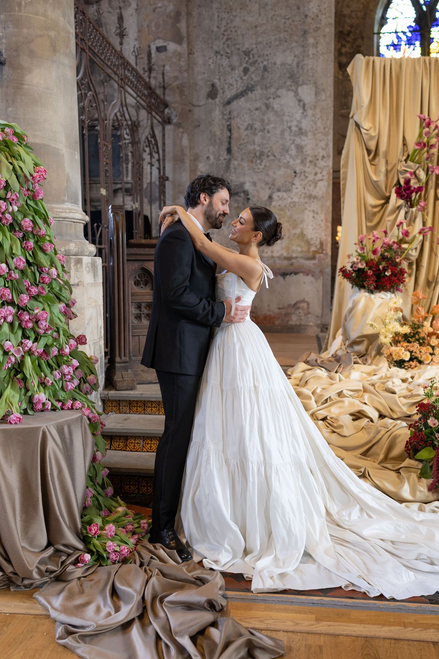 A romantic wedding couple embraces in an ornate church setting decorated with flowing fabrics and pink floral arrangements.