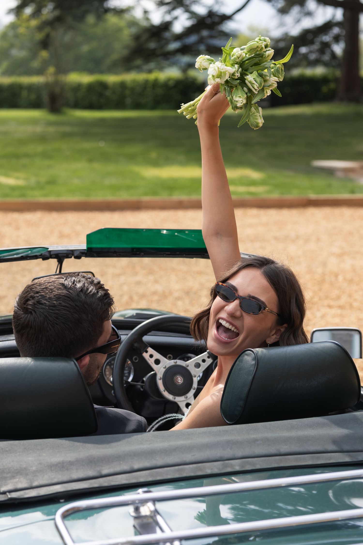 Joyful bride raising bouquet in vintage convertible car with groom, celebrating after wedding ceremony.
