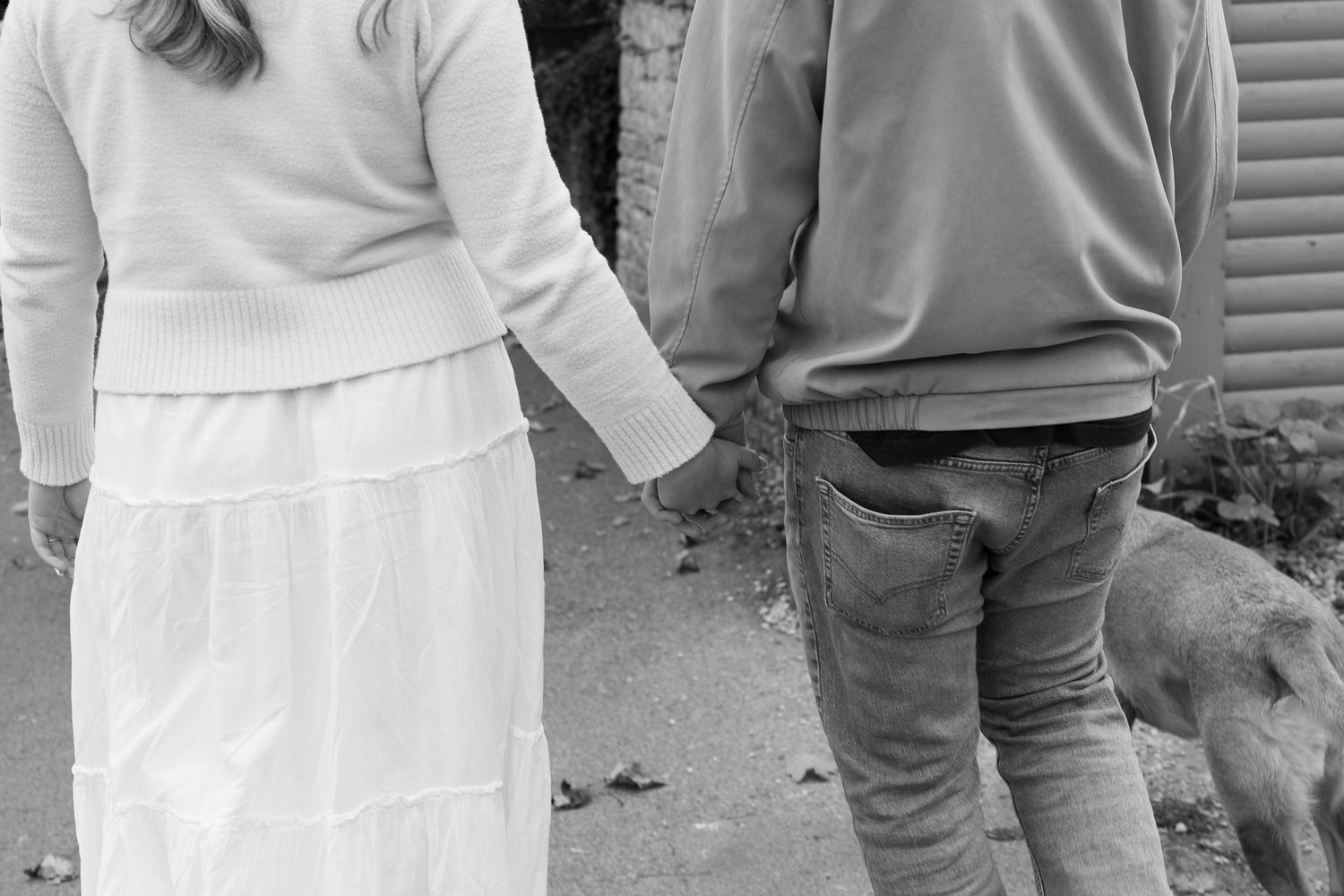 A black and white image of a couple holding hands while walking with their dog.