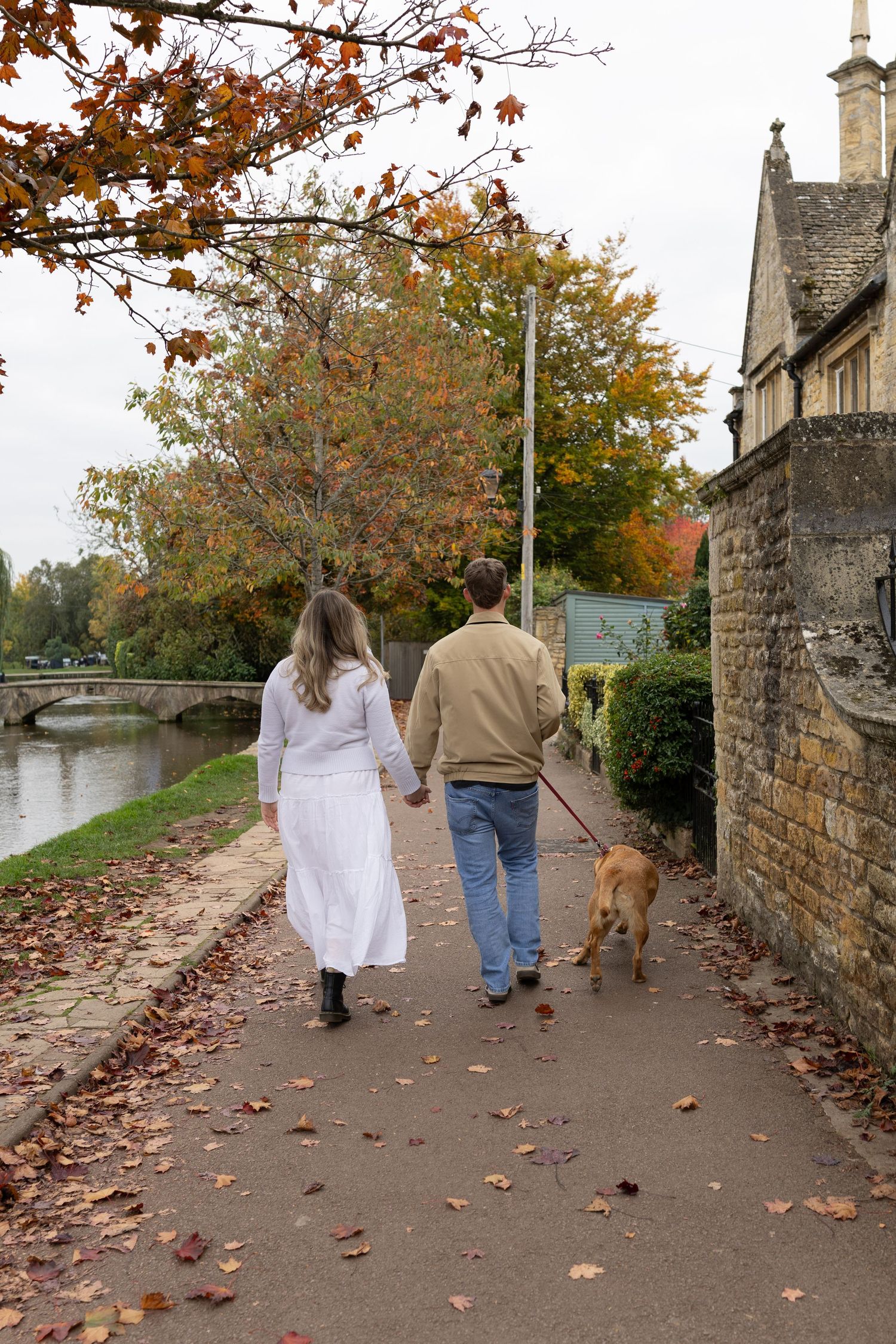 A couple walks their dog along an autumn path beside stone buildings.