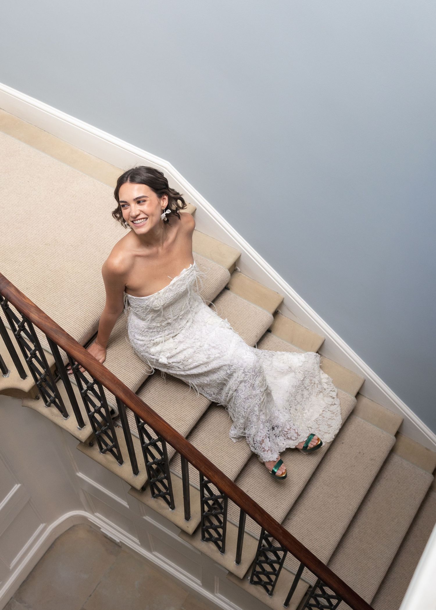 Bride in a strapless lace wedding gown smiling on a carpeted staircase with ornate iron railings.