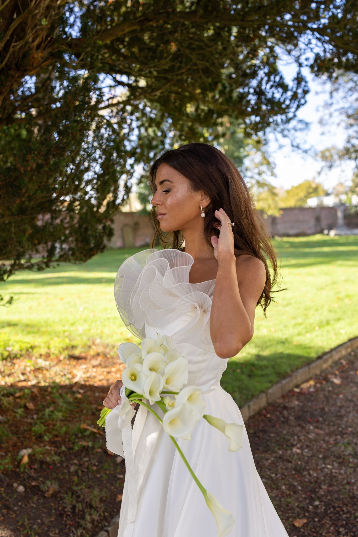 A bride in a flowing white dress poses with white calla lilies in a sunlit outdoor garden setting.