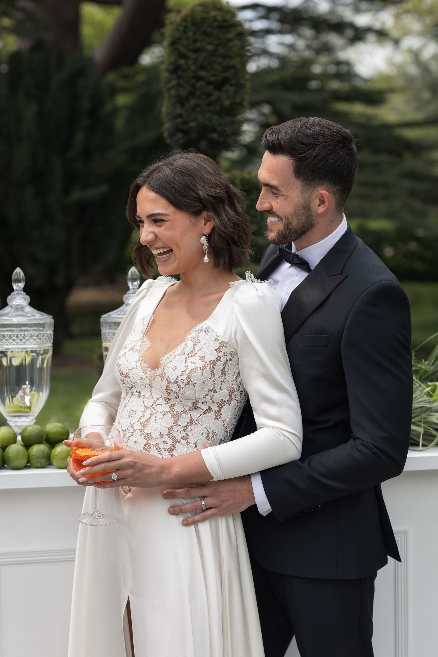 Bride in lace wedding gown laughing with groom in black tuxedo at outdoor wedding reception.