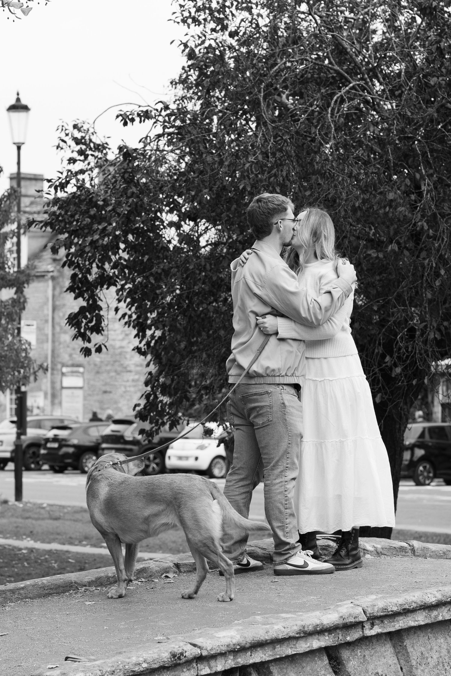 A couple embraces on a stone wall with their dog in black and white.