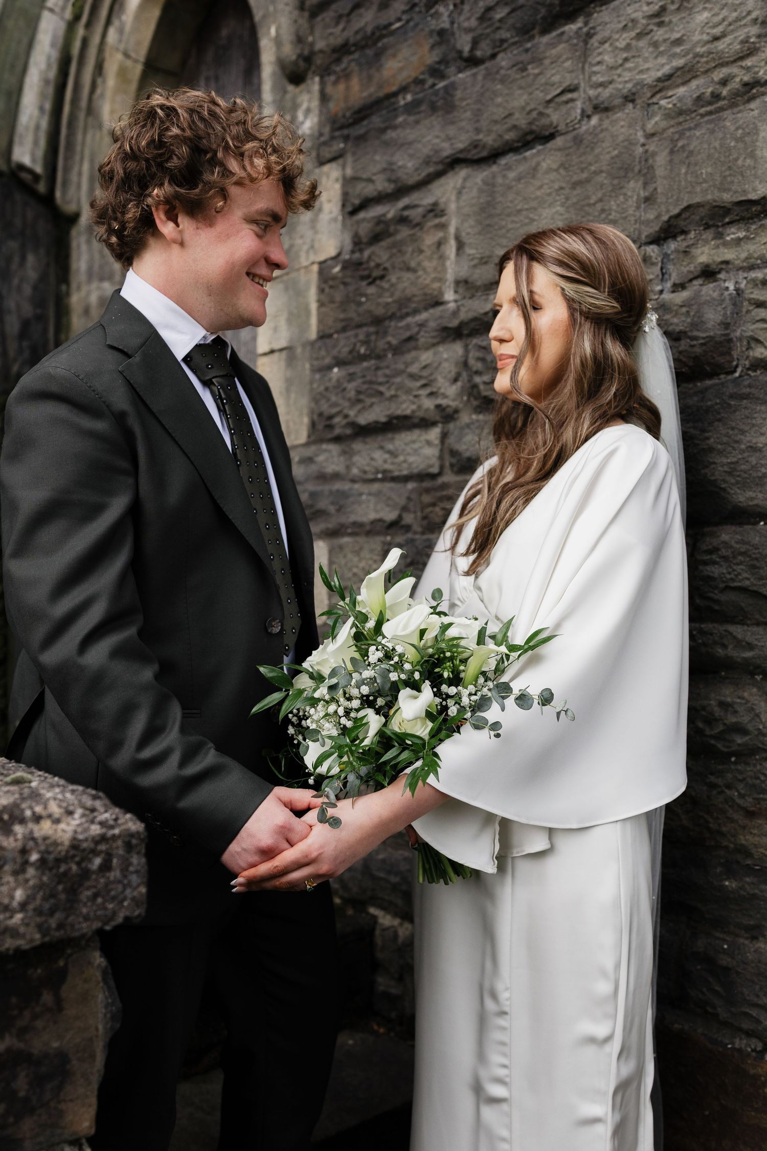A couple shares a tender moment against a stone church wall with white flowers.