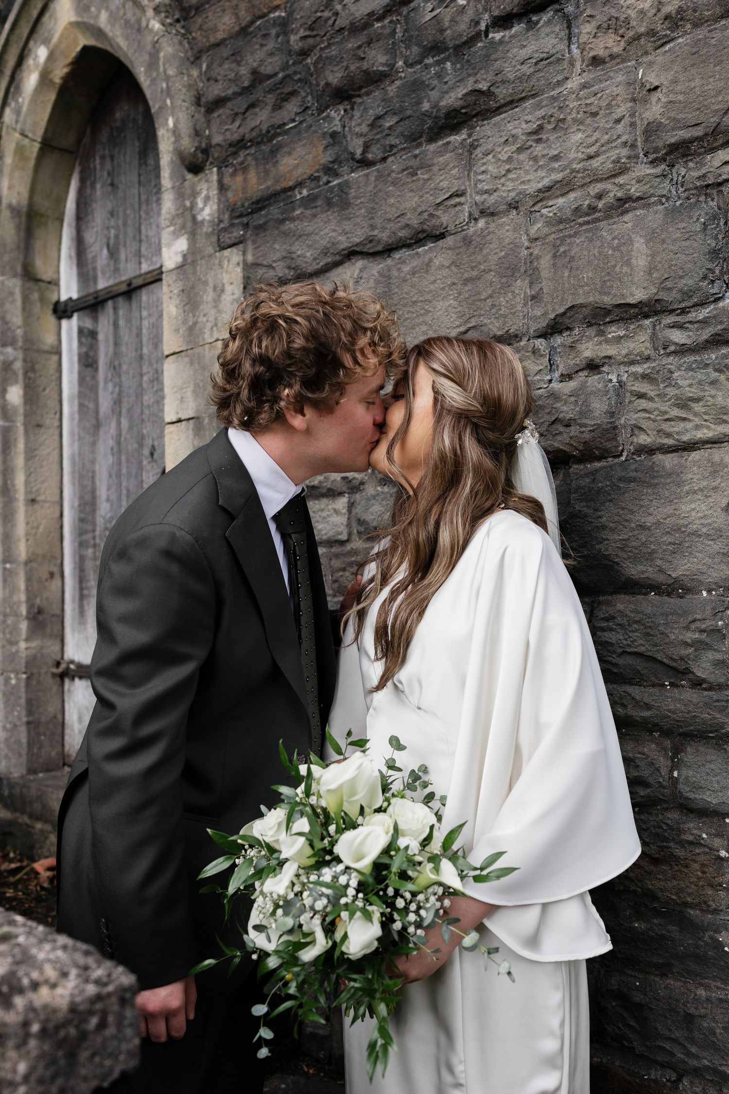 A couple shares an intimate moment against a stone church wall with a white bridal bouquet.