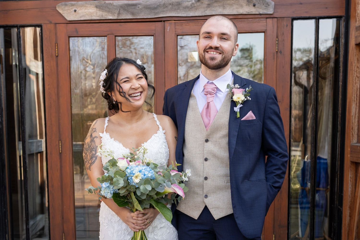 Smiling bride holding blue bouquet stands with groom in navy suit and pink tie outside rustic venue doors.