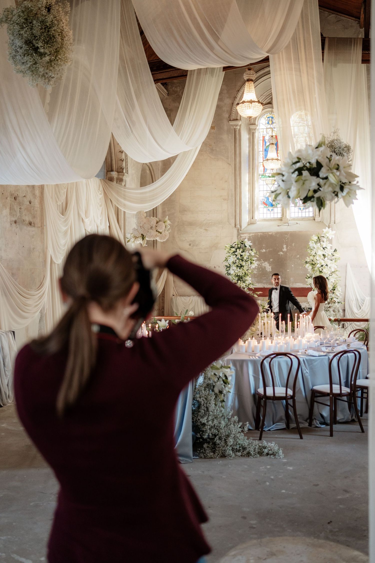 Elegant wedding venue interior with white drapes, chandeliers, and vintage decor with photographer behind the scenes.