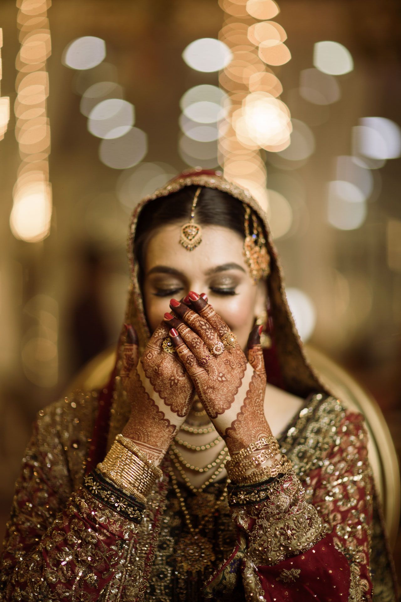 Bride getting ready in a traditional Pakistani bridal room, adorned with gold jewelry and henna-covered hands.