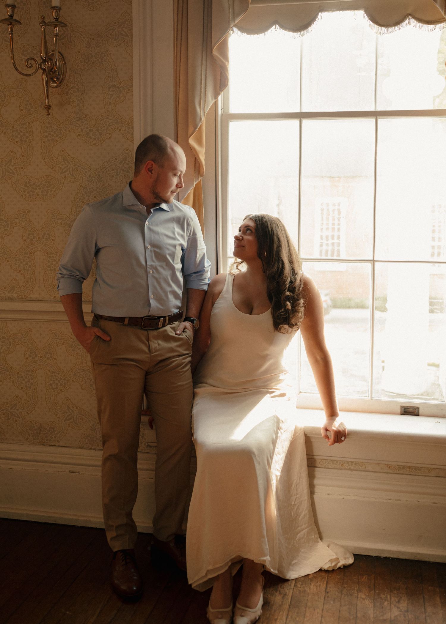 Bride in white dress sitting on window ledge smiling up at groom in sunlit vintage room.