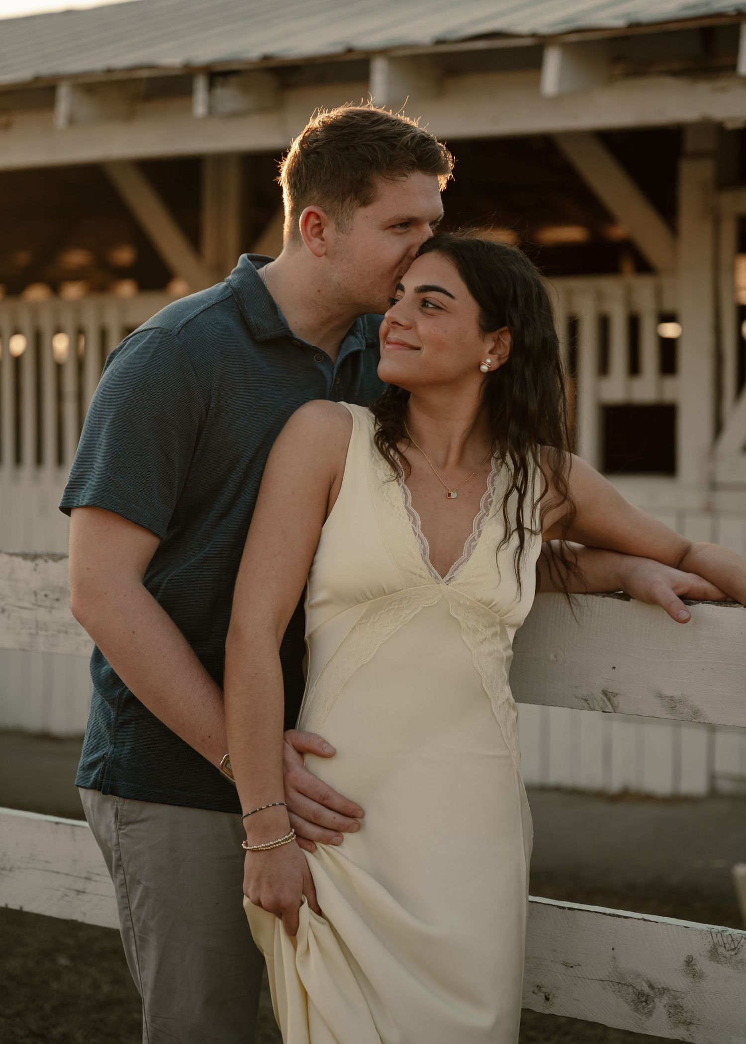Romantic couple embracing near white fence at golden hour with woman in cream slip dress.