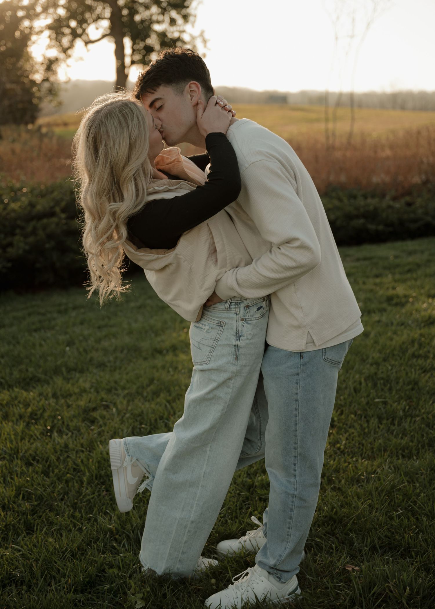 A young couple shares a romantic dip kiss on a grassy lawn during a warm golden sunset.