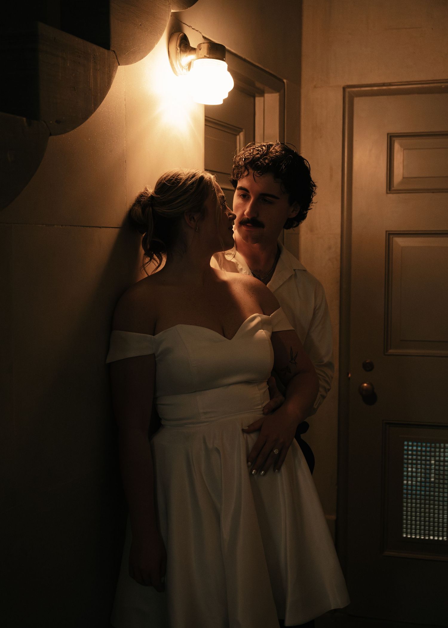 Couple sharing a romantic moment in dim hallway light, woman in white off-shoulder dress, man in white shirt.