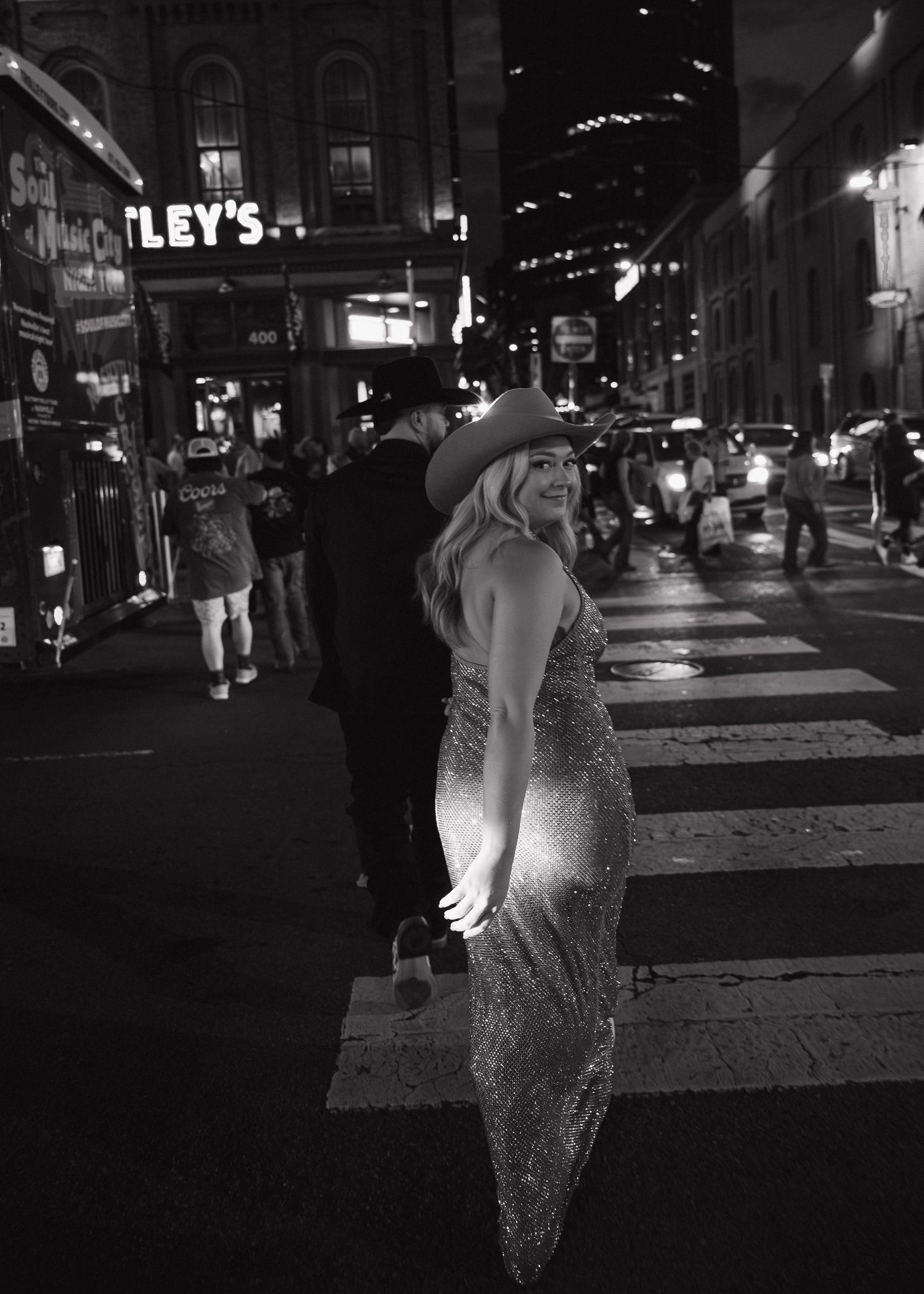 Woman in sequin gown and cowboy hat poses on city street at night in black and white photo.