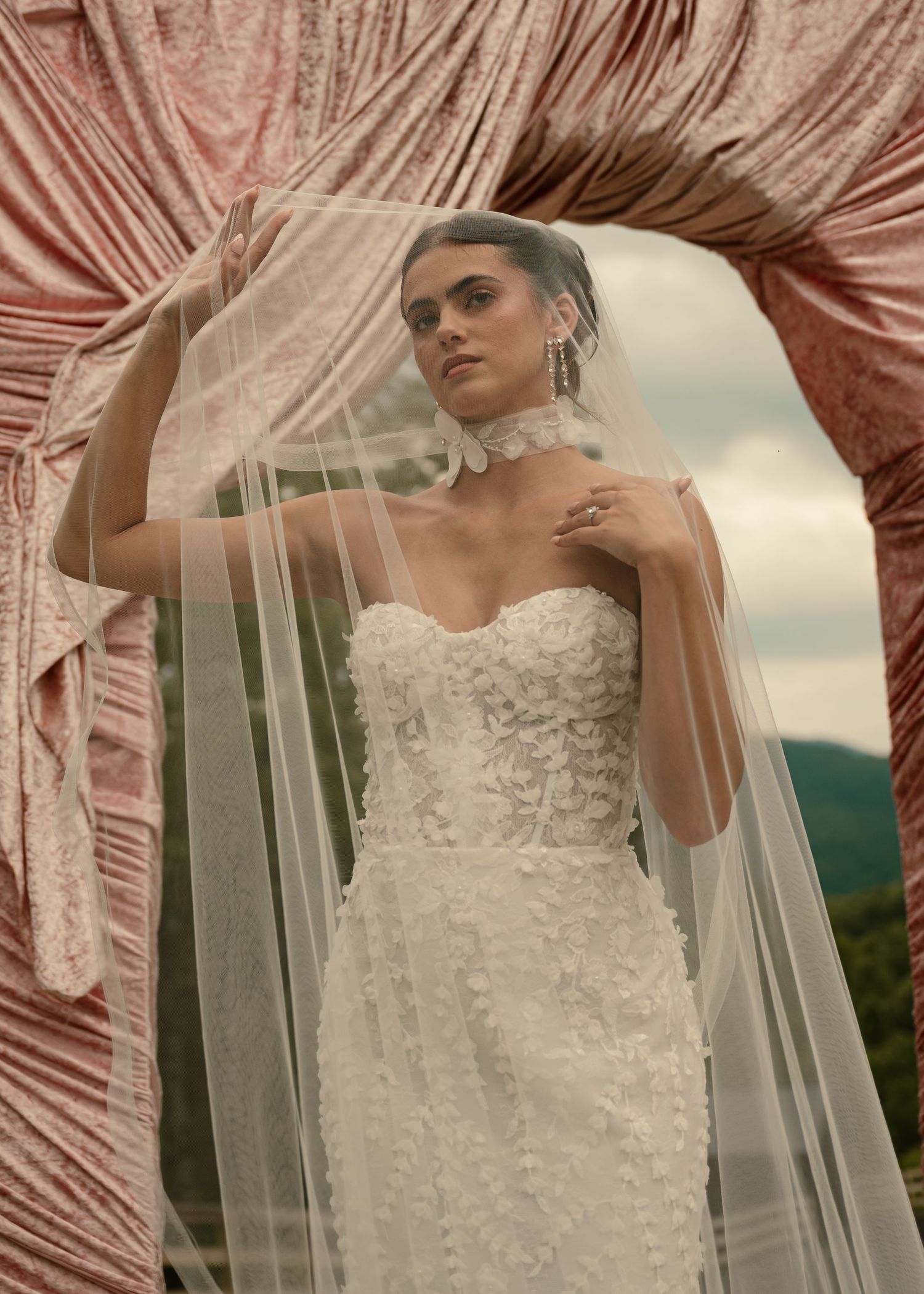 Bride in strapless floral lace wedding gown with tulle veil, posed under draped pink velvet arch outdoors.