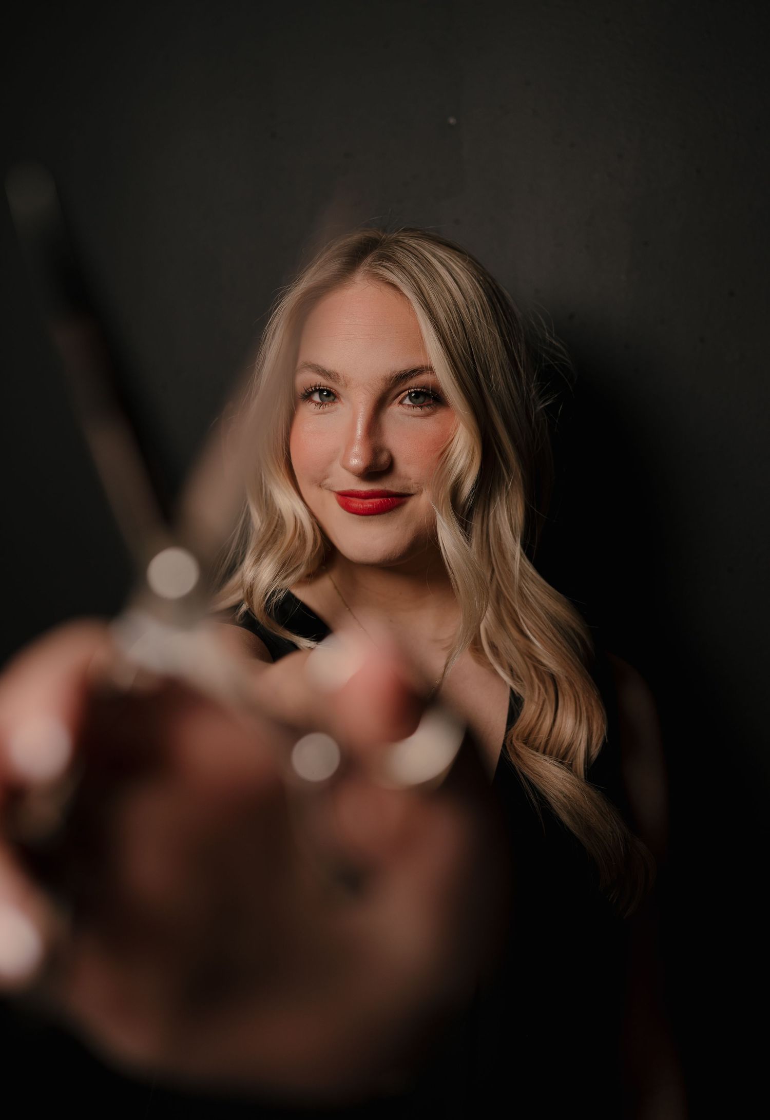 Blonde woman with red lips holding camera lens toward viewer in moody studio portrait.