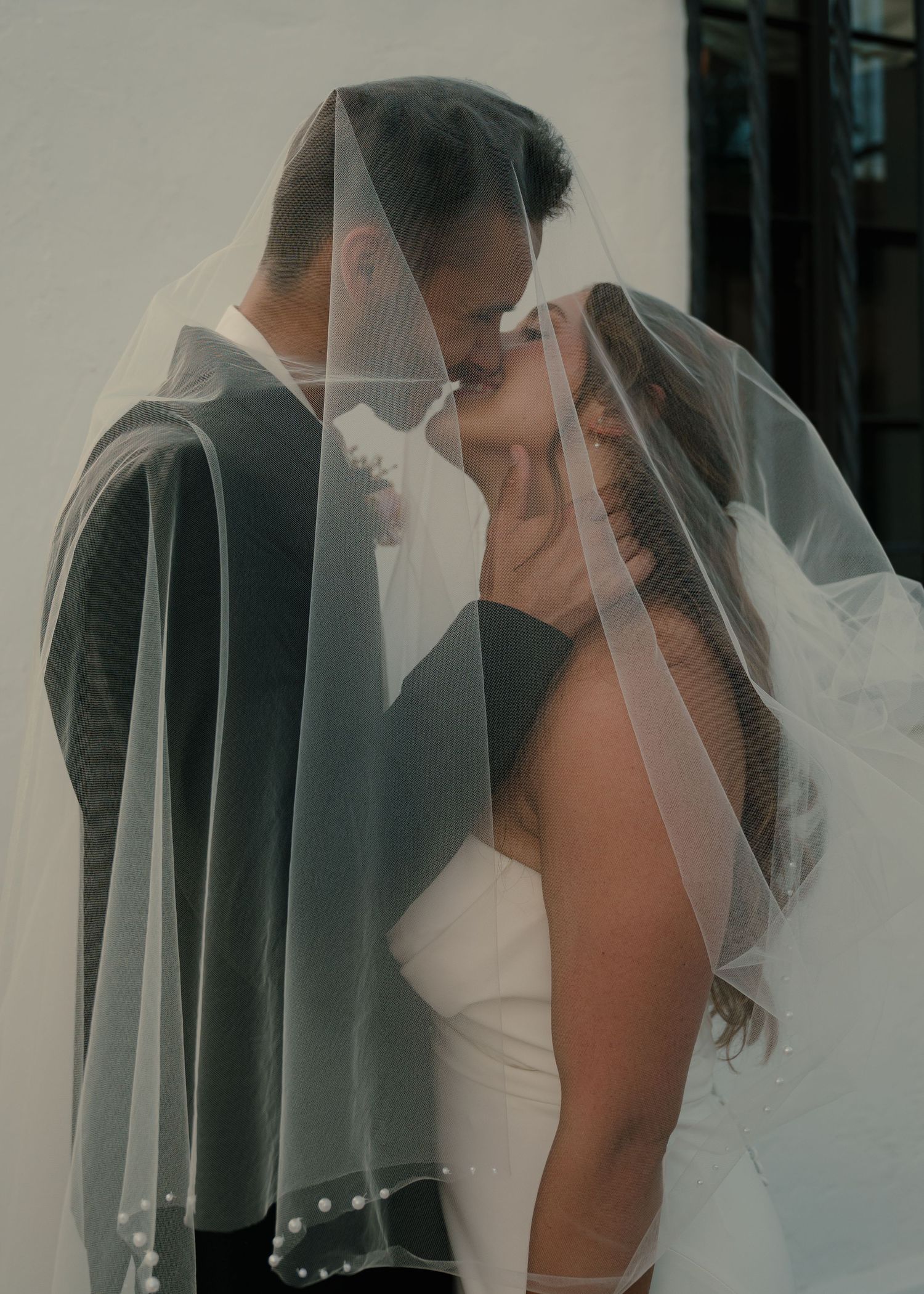Bride and groom sharing romantic kiss under flowing white veil with pearl trim on wedding day.