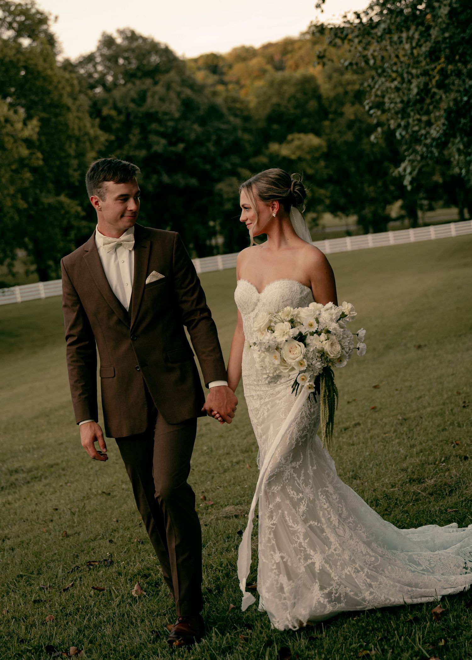 Smiling bride in strapless lace gown and groom in brown suit walk hand in hand on grassy wedding venue. Tennessee Wedding Photographer.