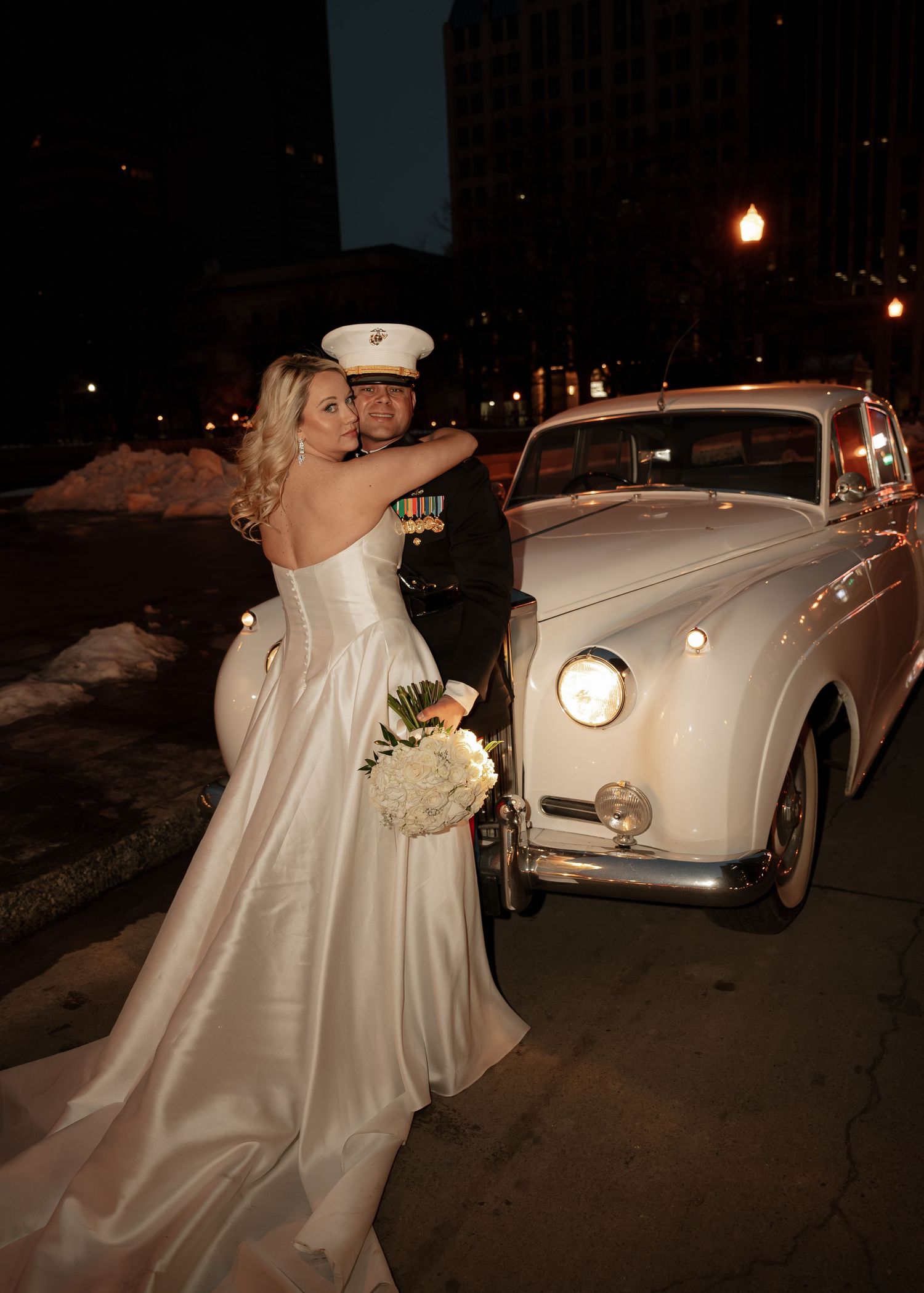 Bride in satin gown hugs Marine groom in dress uniform beside vintage white car at night wedding.