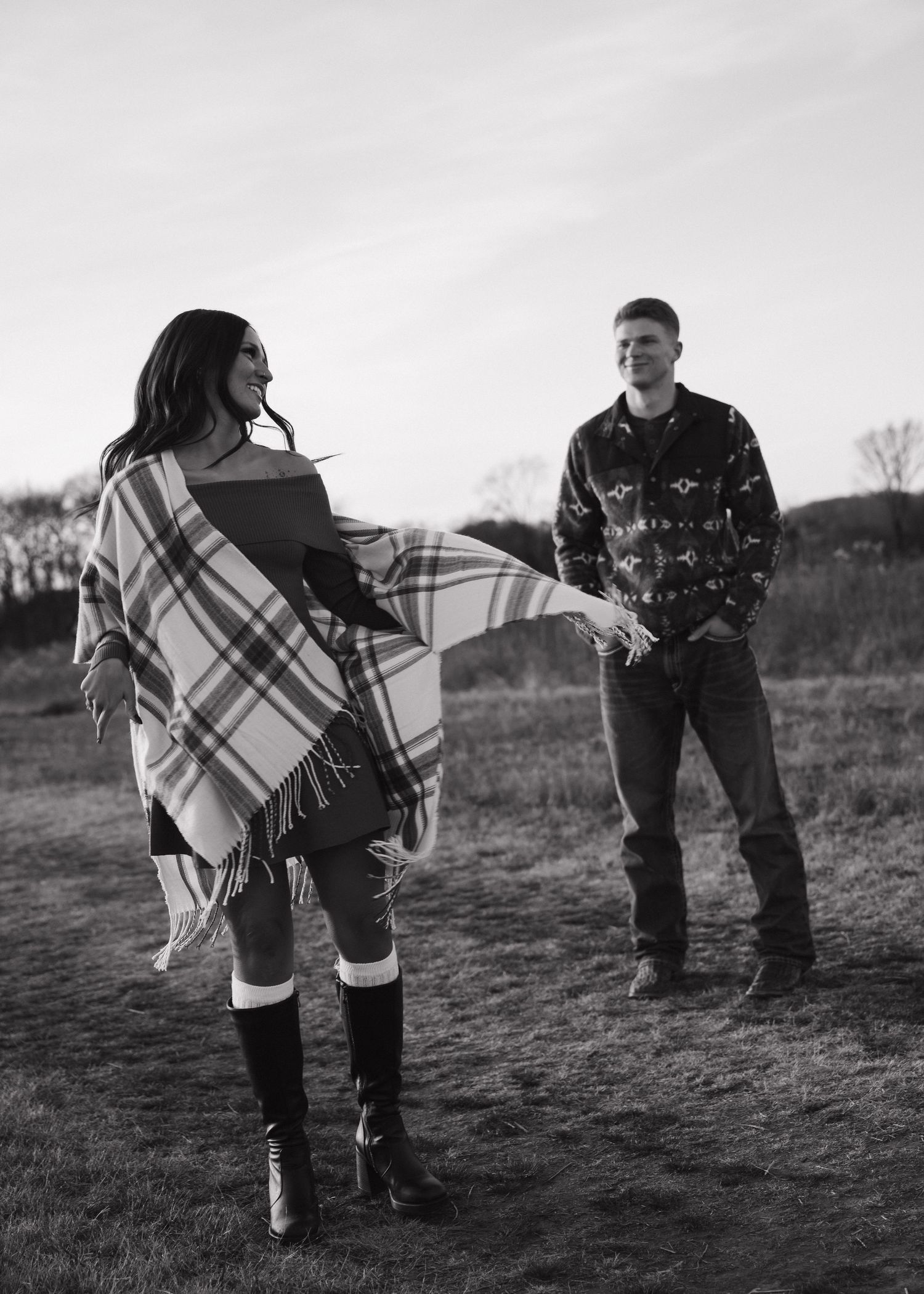 Black and white photo of laughing couple holding hands in an open field, woman twirling in plaid wrap poncho.