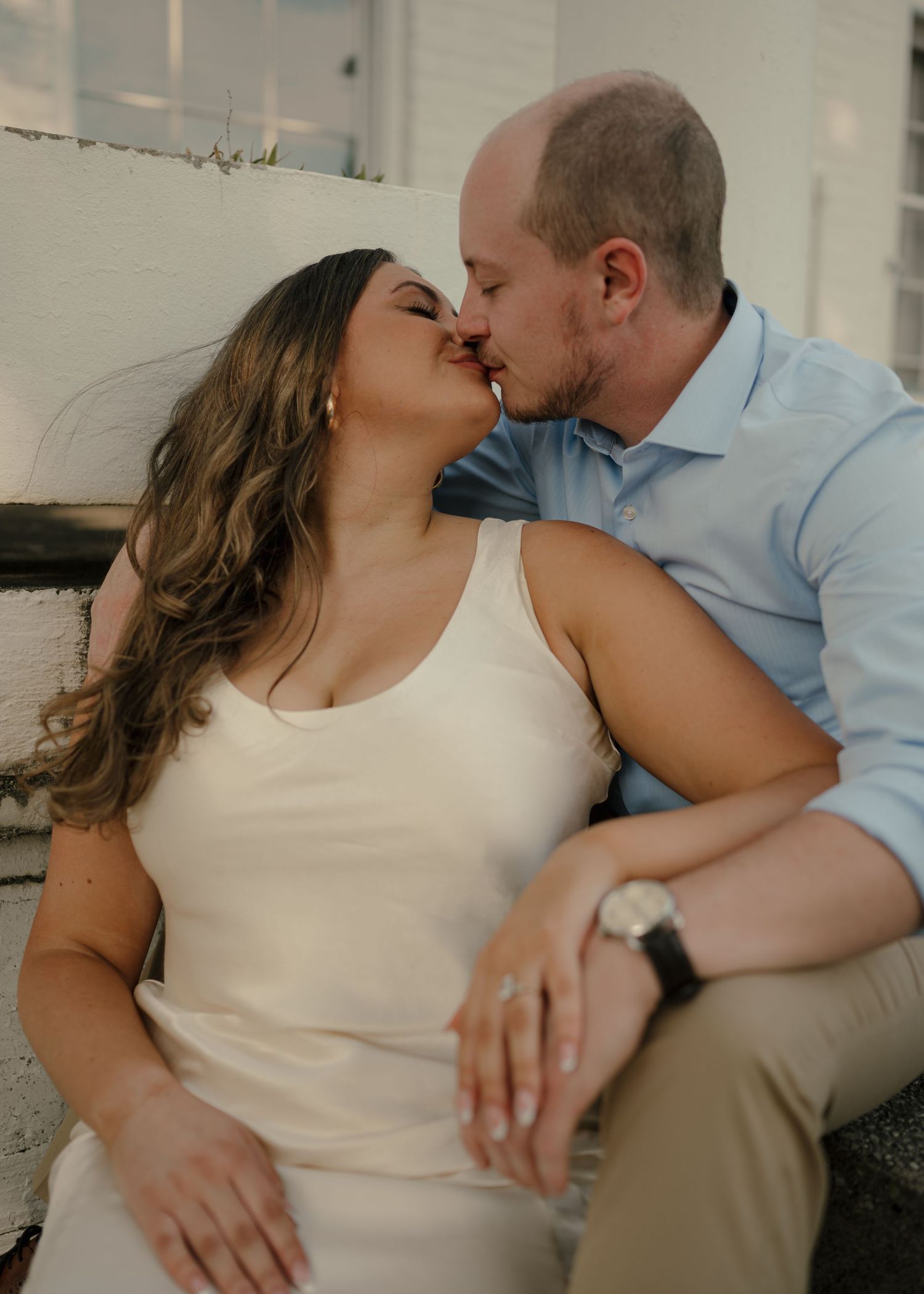 Couple sharing a romantic kiss while sitting outdoors against a white brick wall.