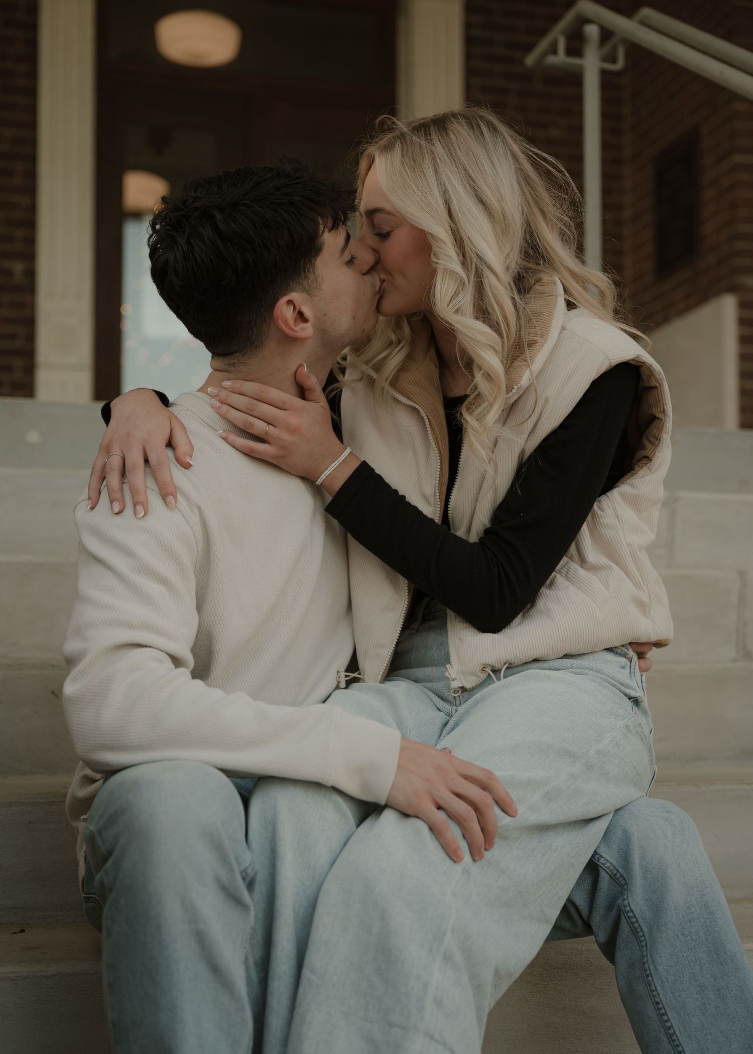 A couple shares a kiss while seated on outdoor steps, dressed casually in light denim and neutral tones.