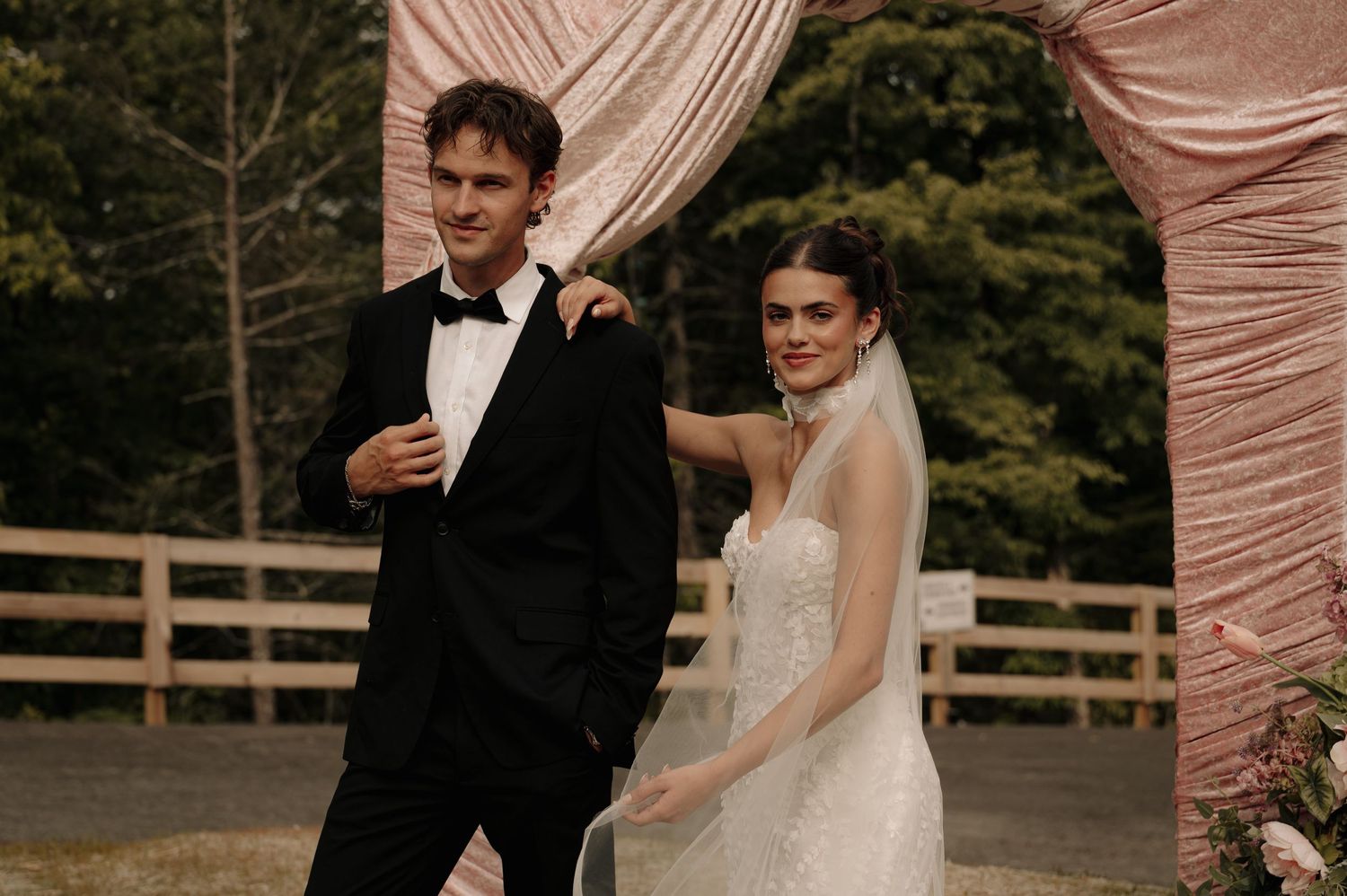 Bride in white gown and groom in black tuxedo stand together outdoors under pink draped wedding arch.