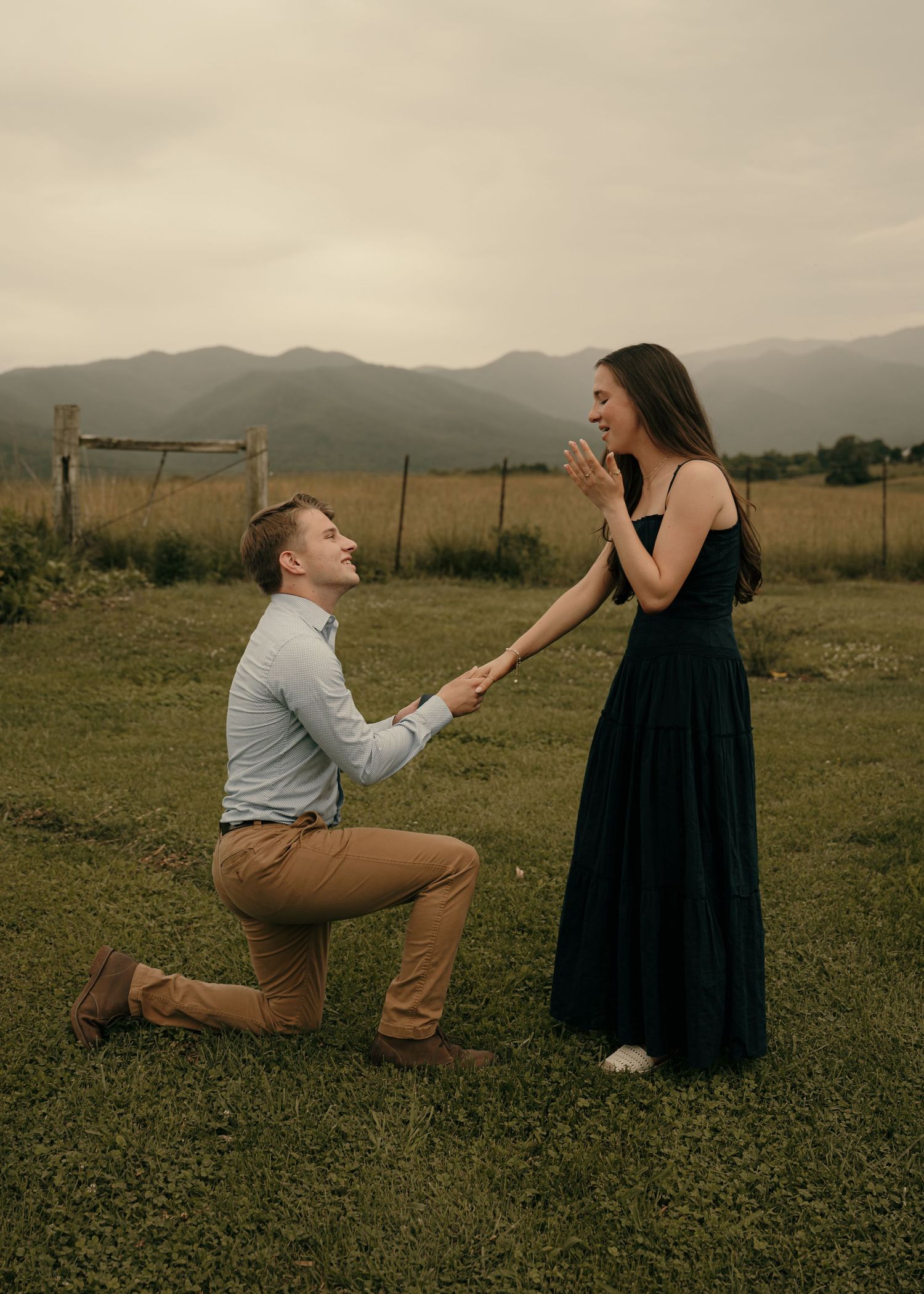Man proposing to woman in a mountain meadow, she reacts with joy and surprise.
