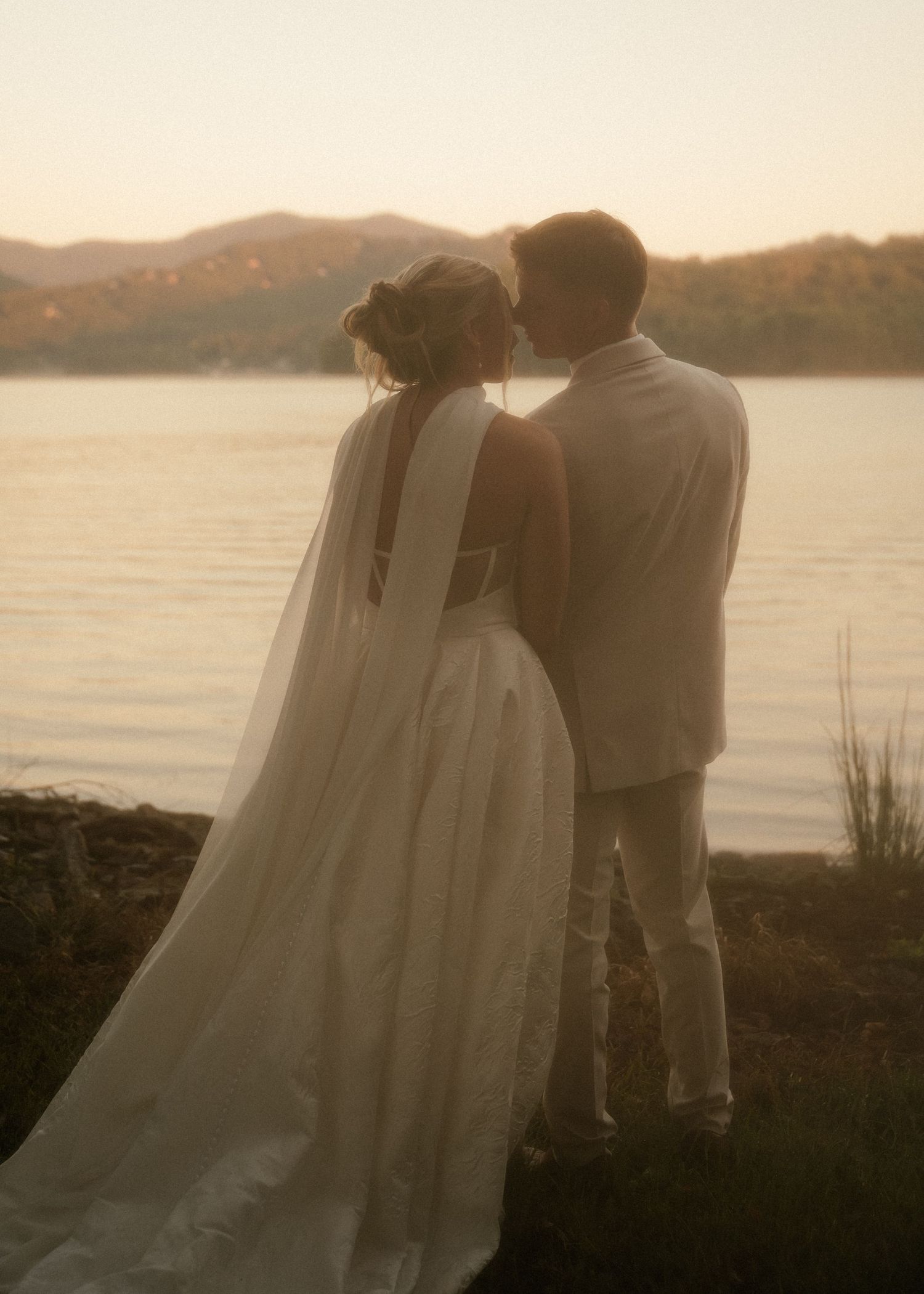 Bride and groom sharing romantic moment by serene lake with mountain backdrop at sunset.