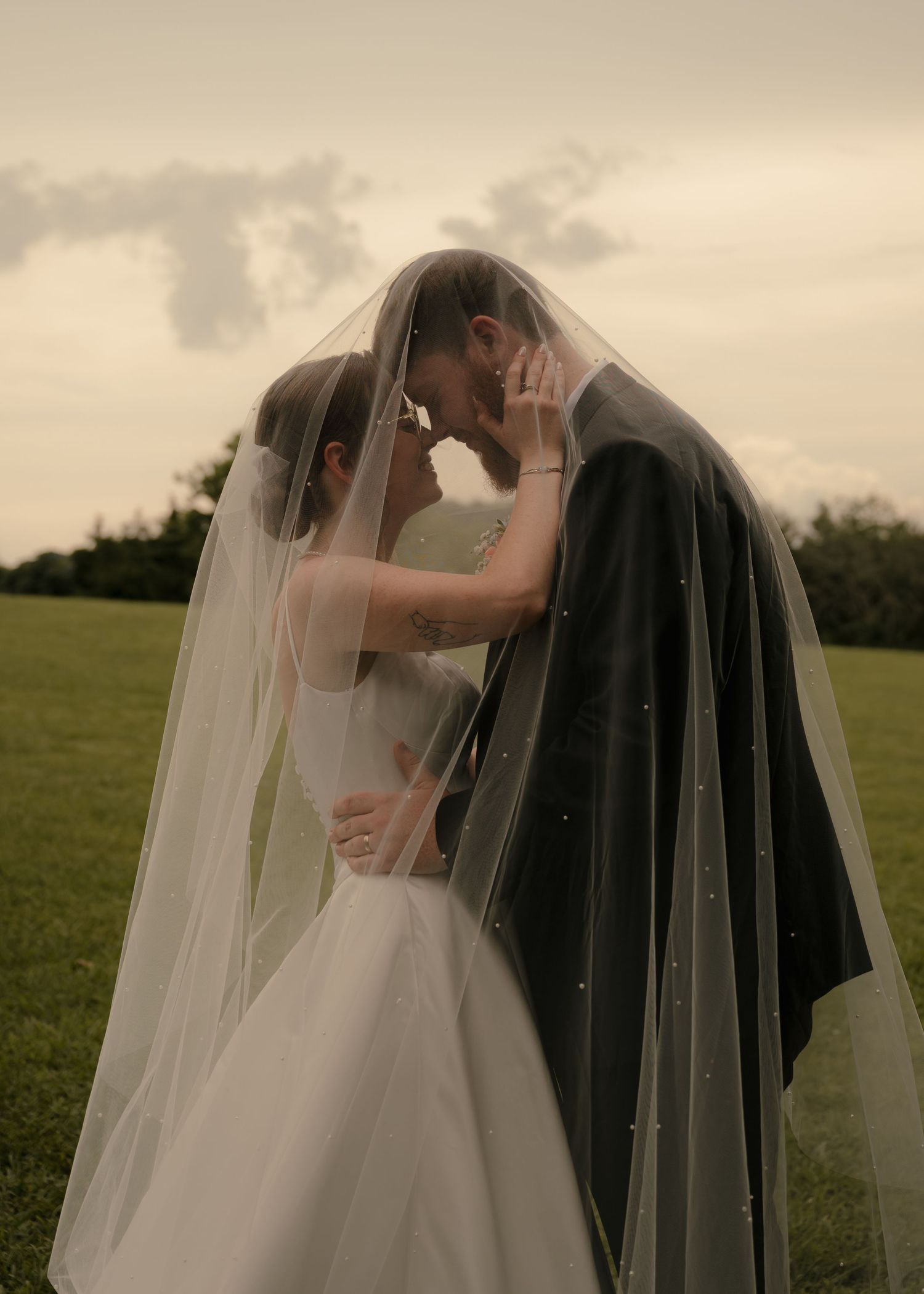 Romantic wedding portrait of couple touching foreheads under flowing pearl wedding veil outdoors.