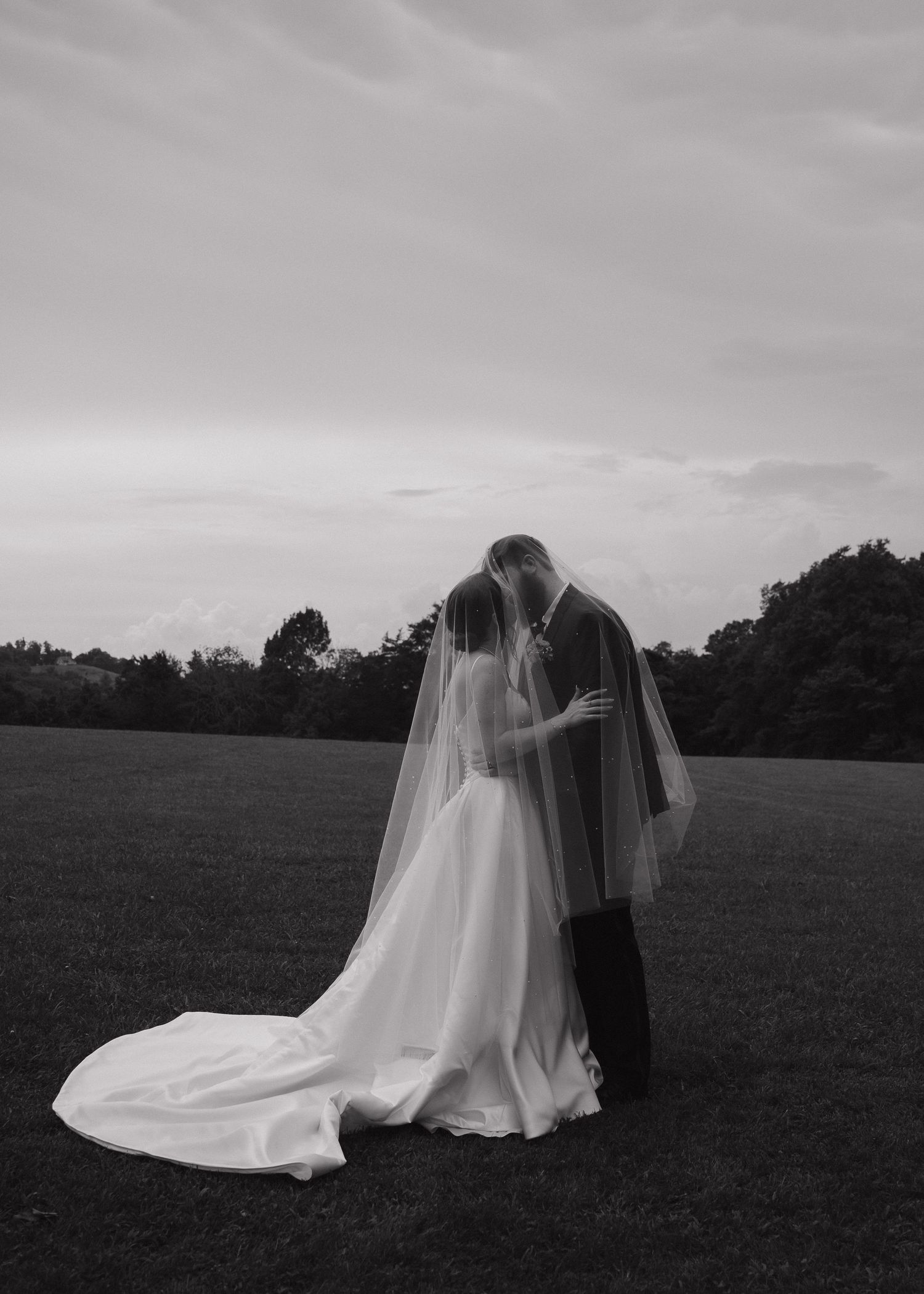 Black and white photo of bride and groom kissing in open field, wedding veil flowing behind them.