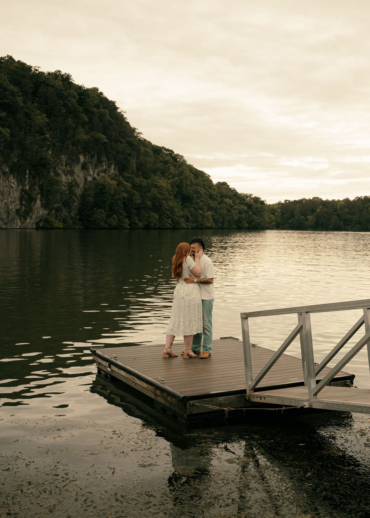 Couple embracing on a wooden dock over a calm lake at dusk, surrounded by lush green hills.