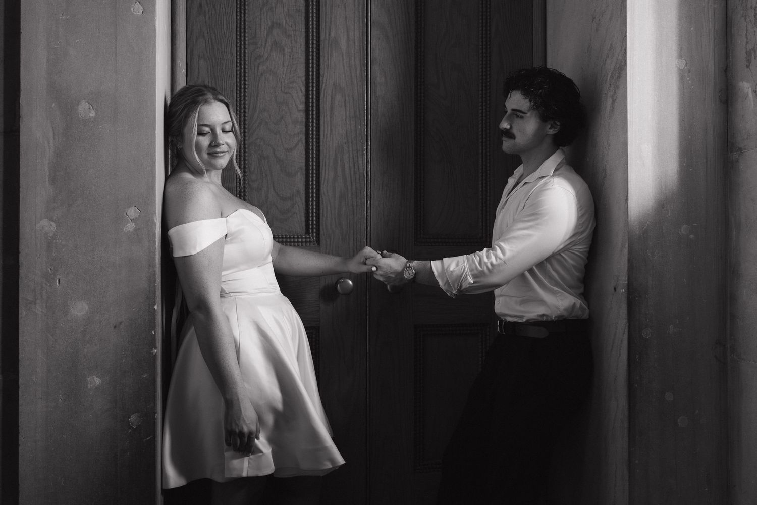 Black and white photo of a couple holding hands in a doorway, woman in white dress smiling at man in white shirt.