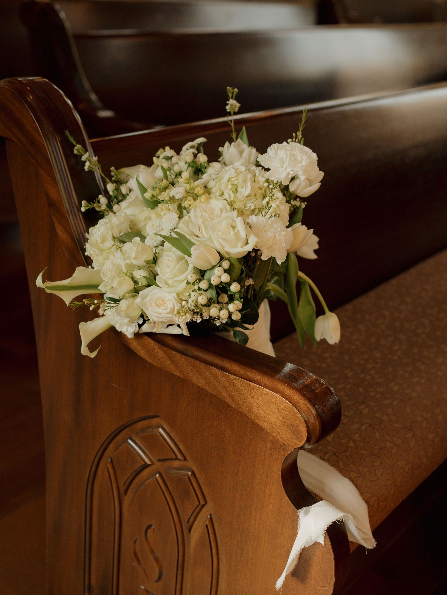 White floral wedding bouquet with roses and tulips adorning a wooden church pew end.