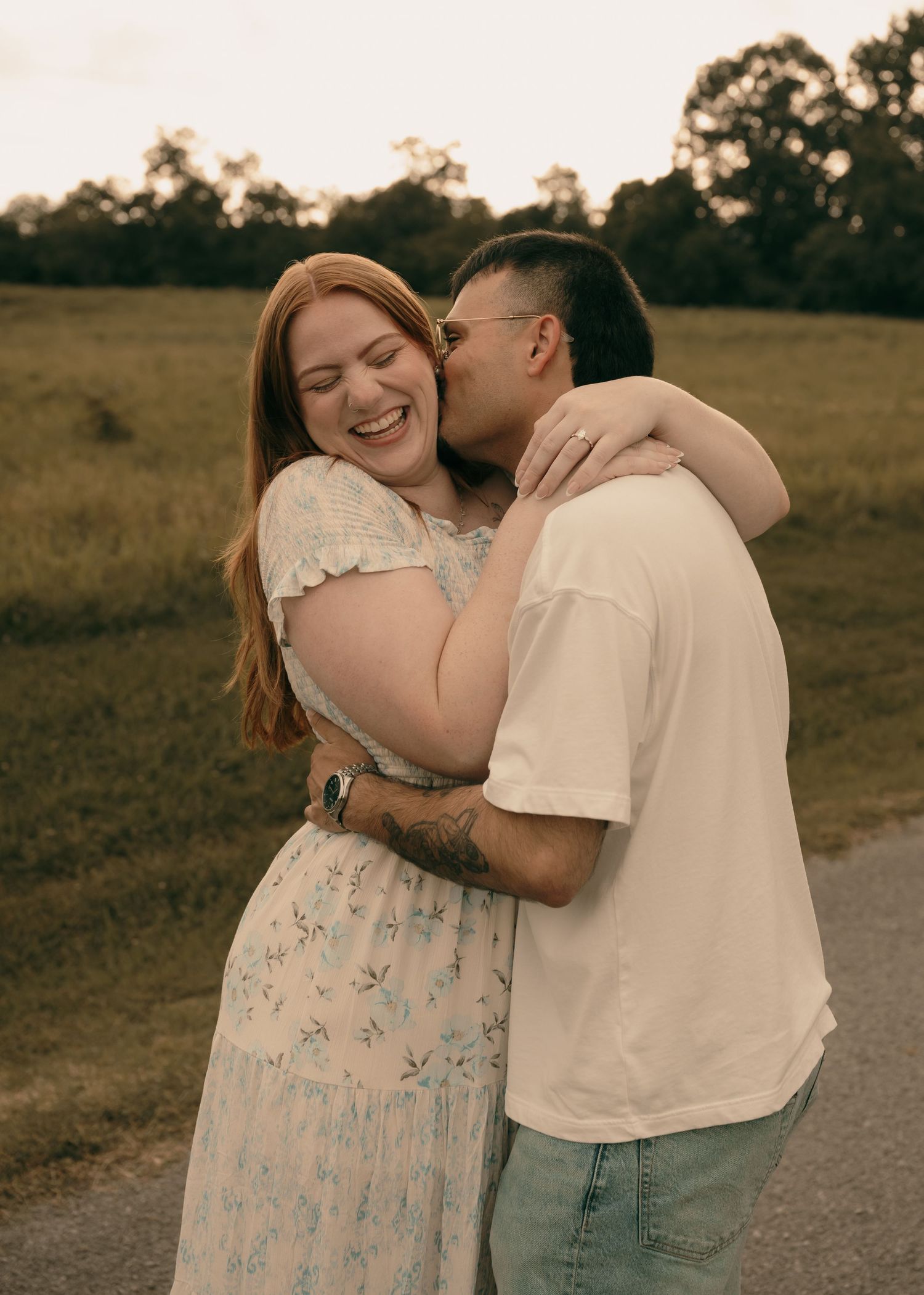 Happy couple embracing outdoors, woman in floral dress laughing as man kisses her cheek in a green field.
