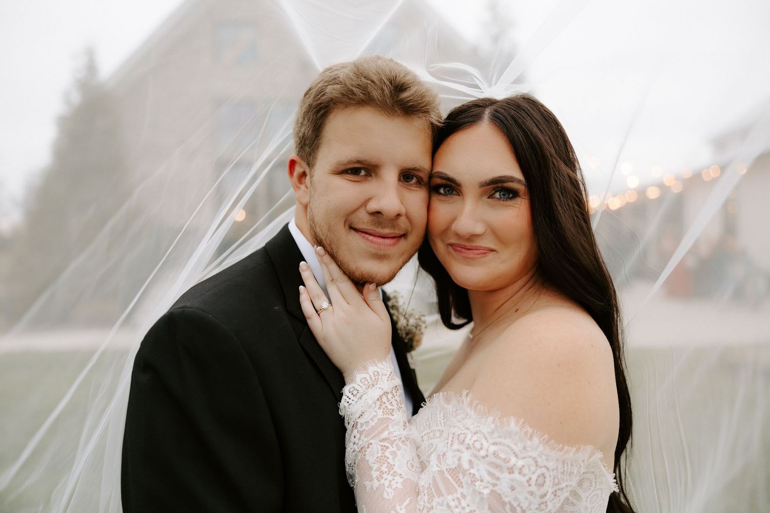 A bride and groom smile together under a flowing veil on a misty wedding day.