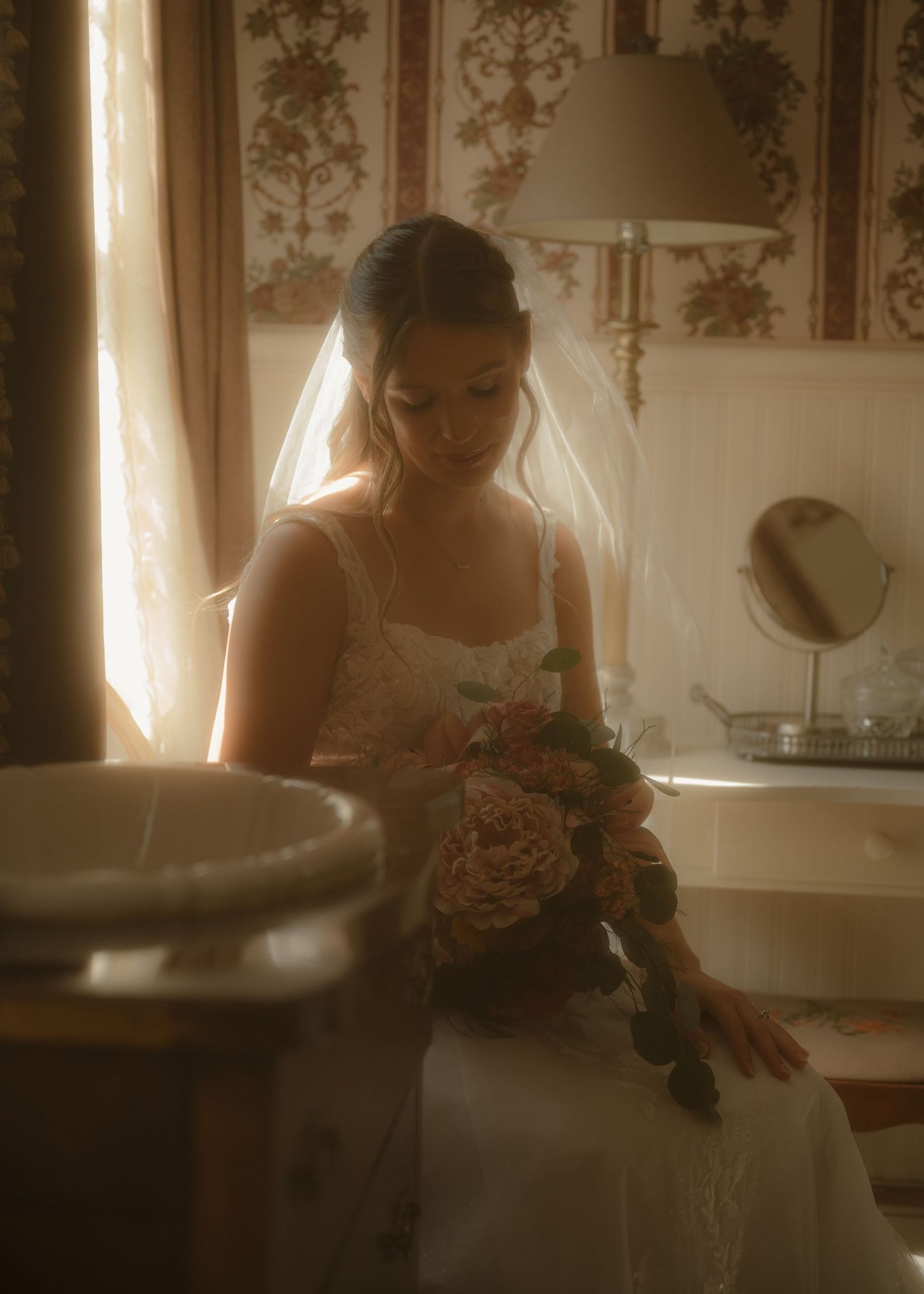 Bride in white gown and veil sitting by window in vintage room, gazing at her bouquet of blush flowers.