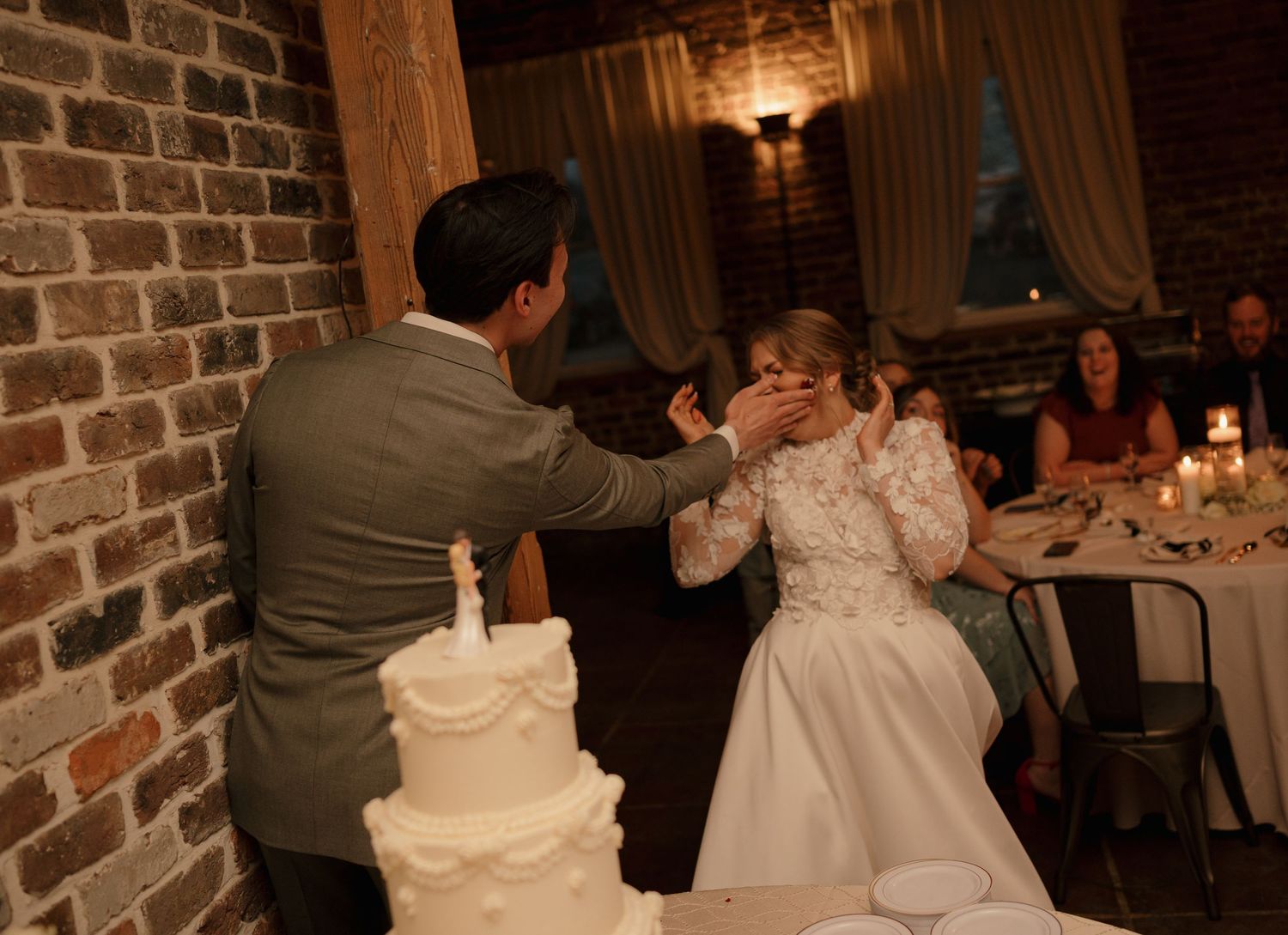 Groom feeding wedding cake to bride as guests laugh at candlelit rustic brick reception venue.