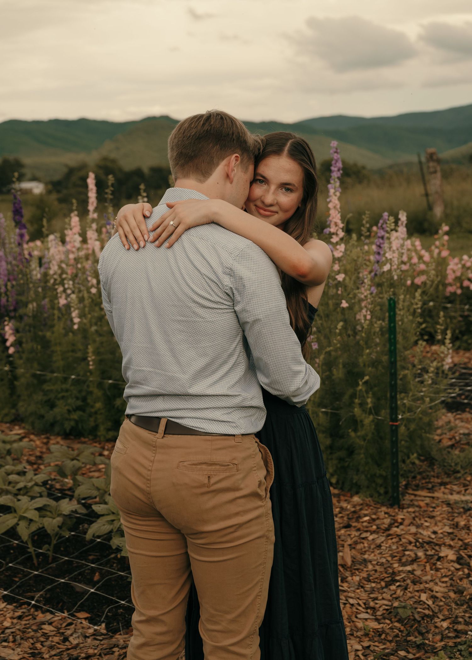 Couple embracing in a flower garden with mountains in the background, woman smiling over mans shoulder.