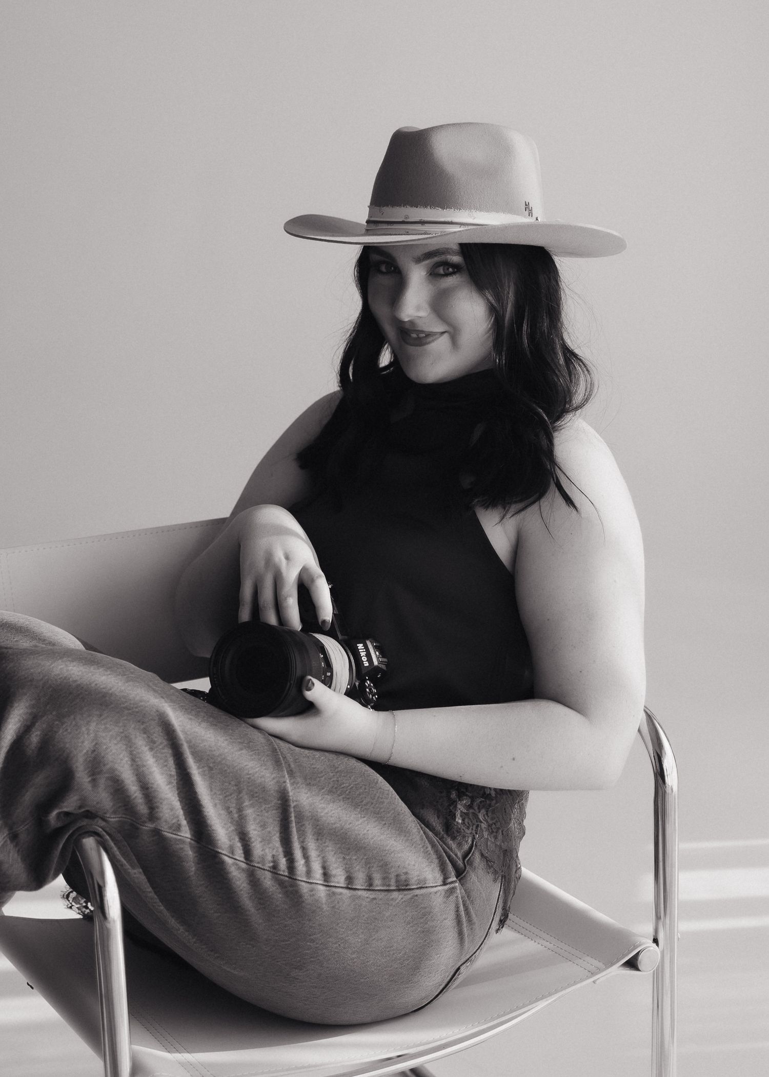 Black and white portrait of smiling woman in hat sitting in chair holding professional camera.