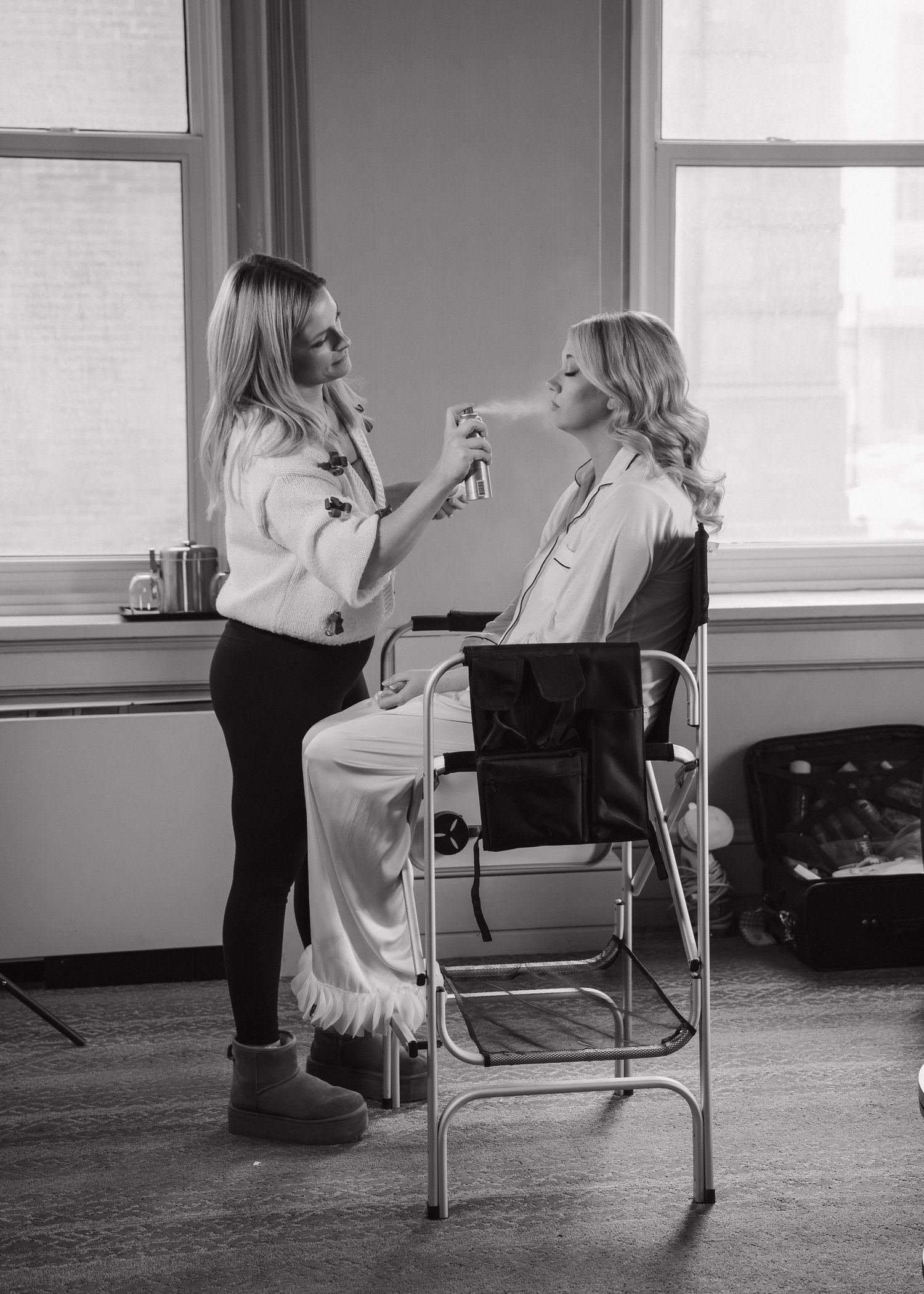Black and white photo of makeup artist applying cosmetics to a bride in a bright windowed room.