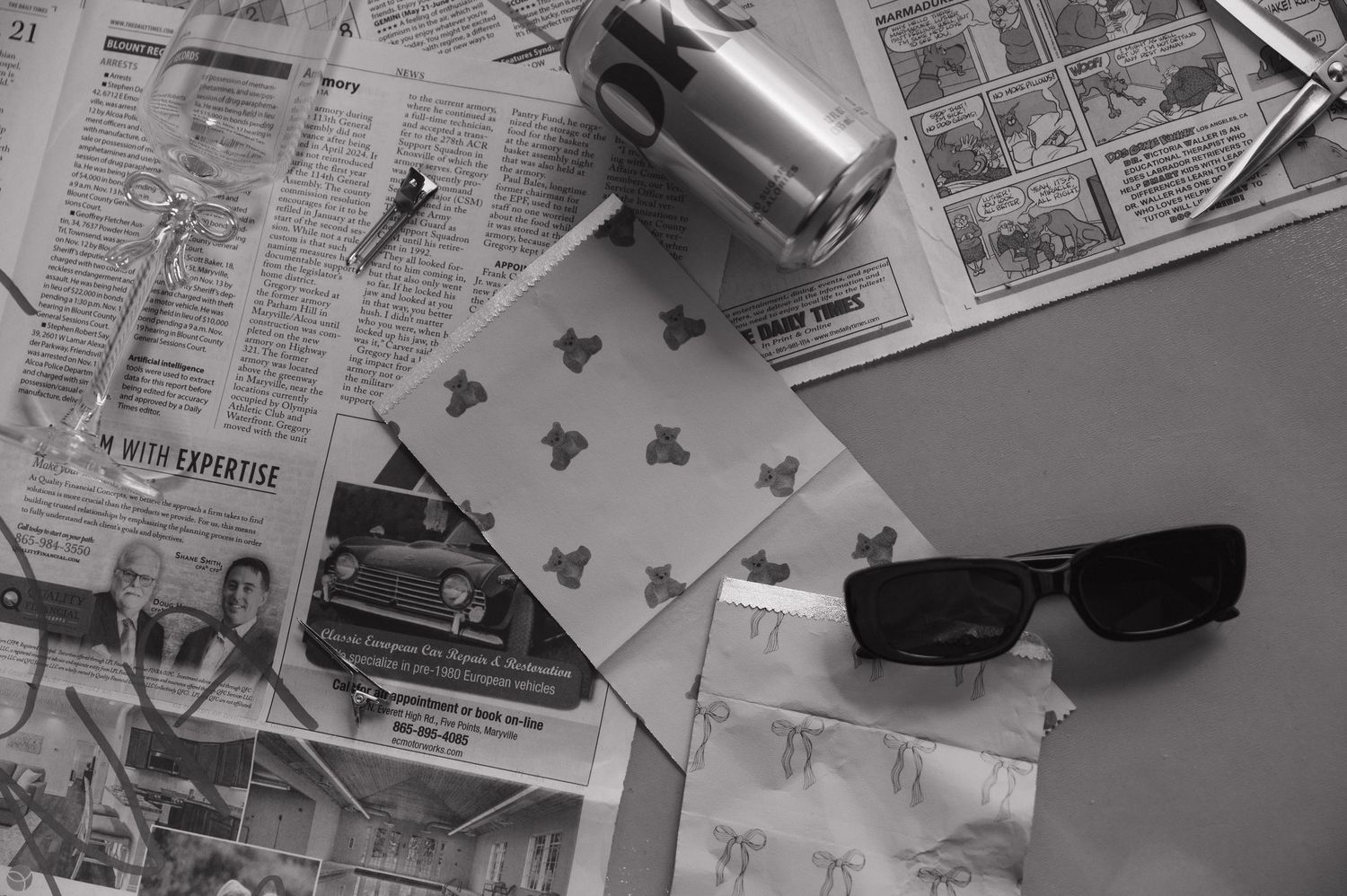 Black and white flat lay of sunglasses, newspaper, playing card, and camera on a desk.